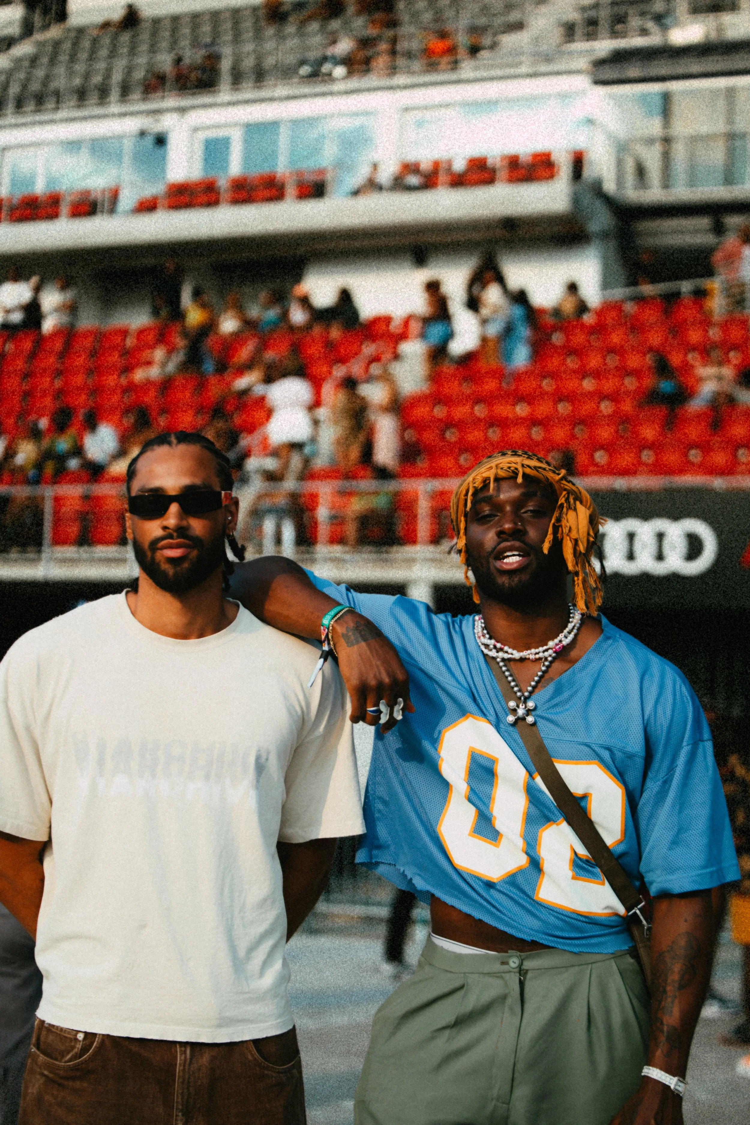 Two men posing in a stadium with red seats in the background, one wearing a white shirt and sunglasses, the other wearing a cropped blue football jersey and accessories.