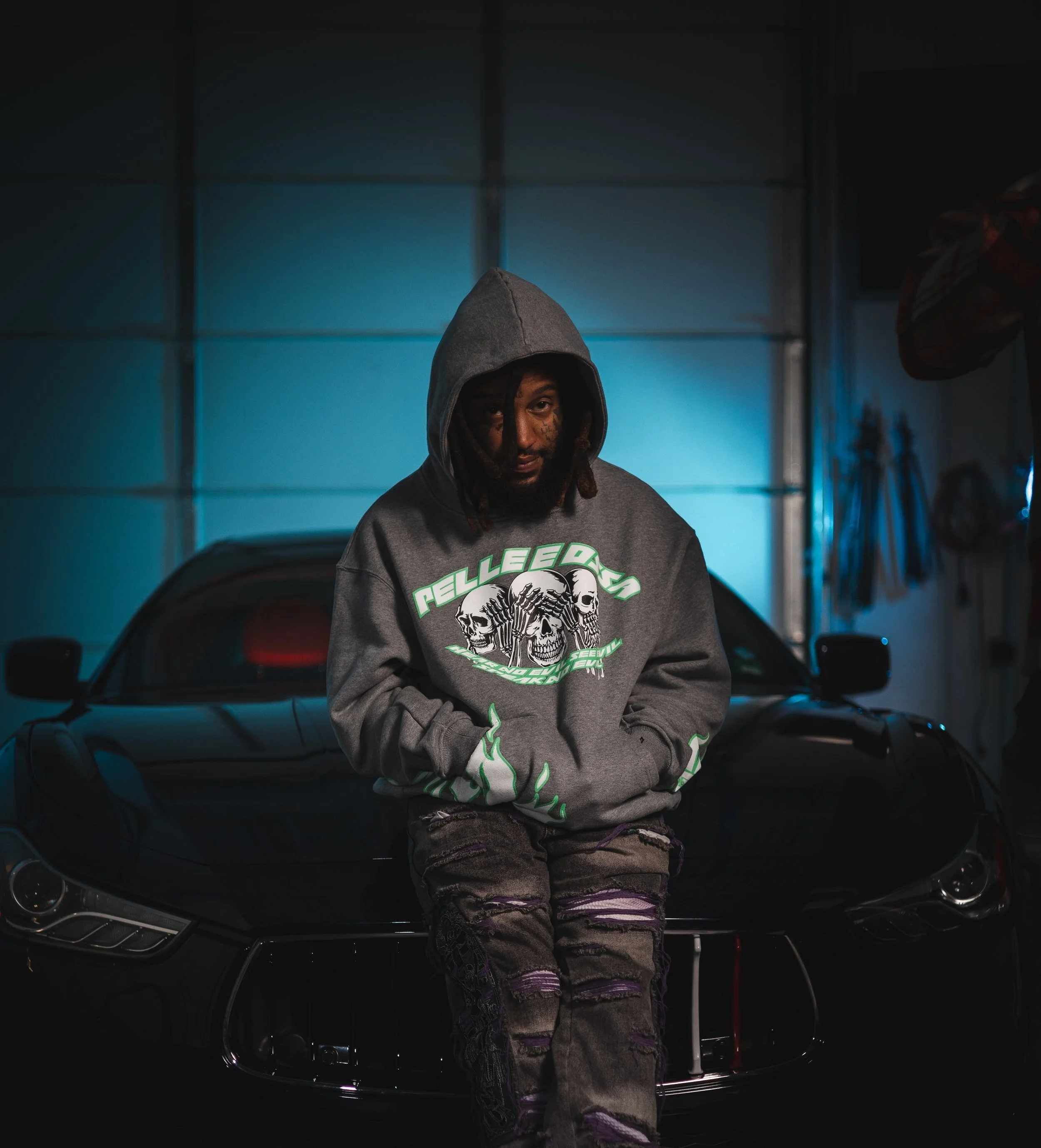 Person in a hooded sweatshirt sitting on a sleek black car in a dimly lit garage.