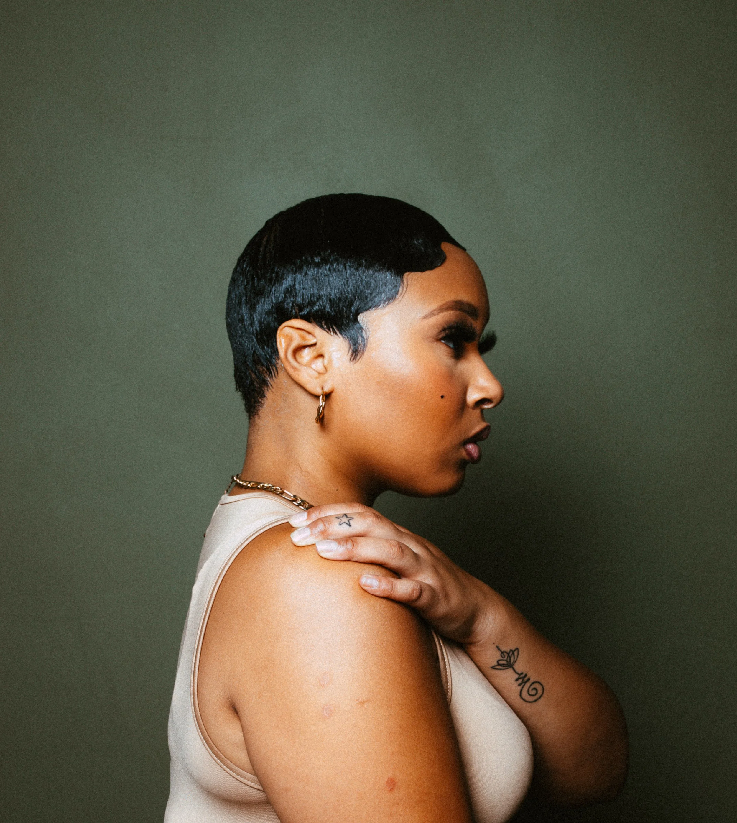 Profile of a person with short, black hair wearing a sleeveless top and gold earring, looking to the left against a green background. They have tattoos on their arm and shoulder.