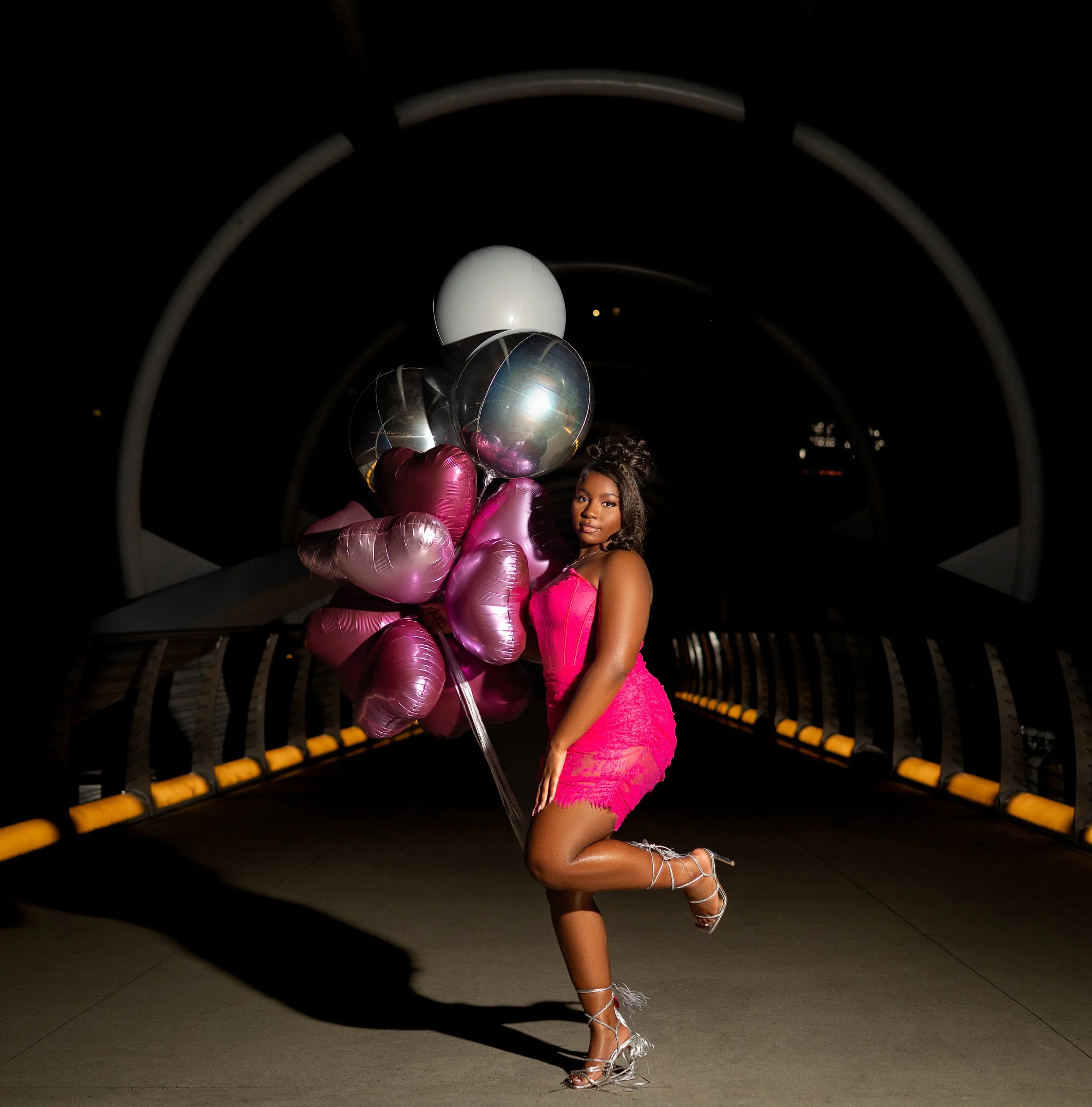 Woman in a bright pink dress holding a bouquet of shiny pink and silver balloons, posing on a dimly lit bridge at night.