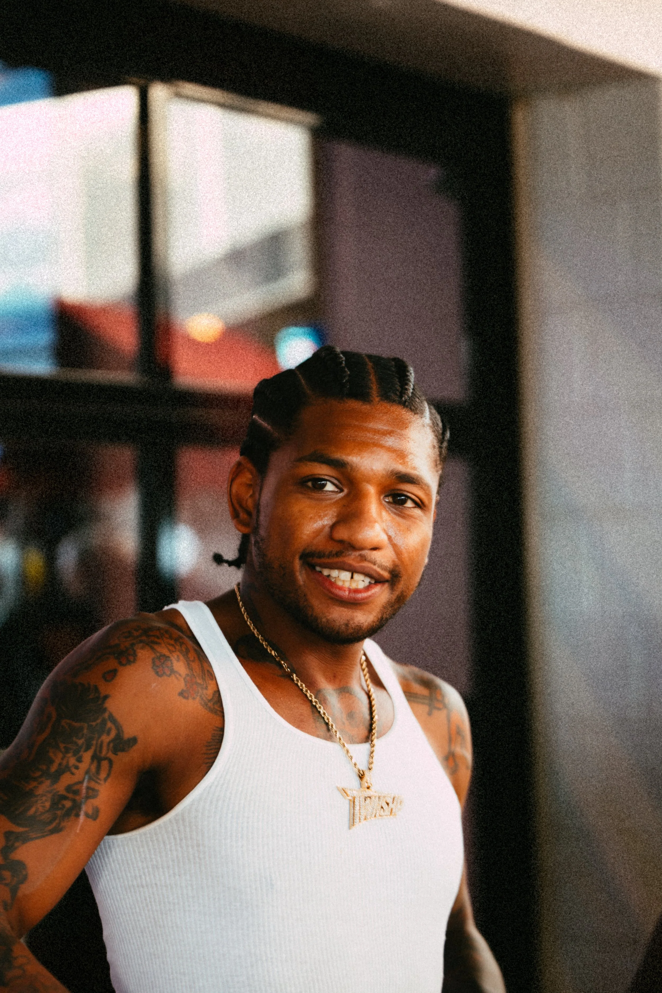 Smiling man with braided hair and tattoos wearing a white tank top and gold necklace, standing indoors.