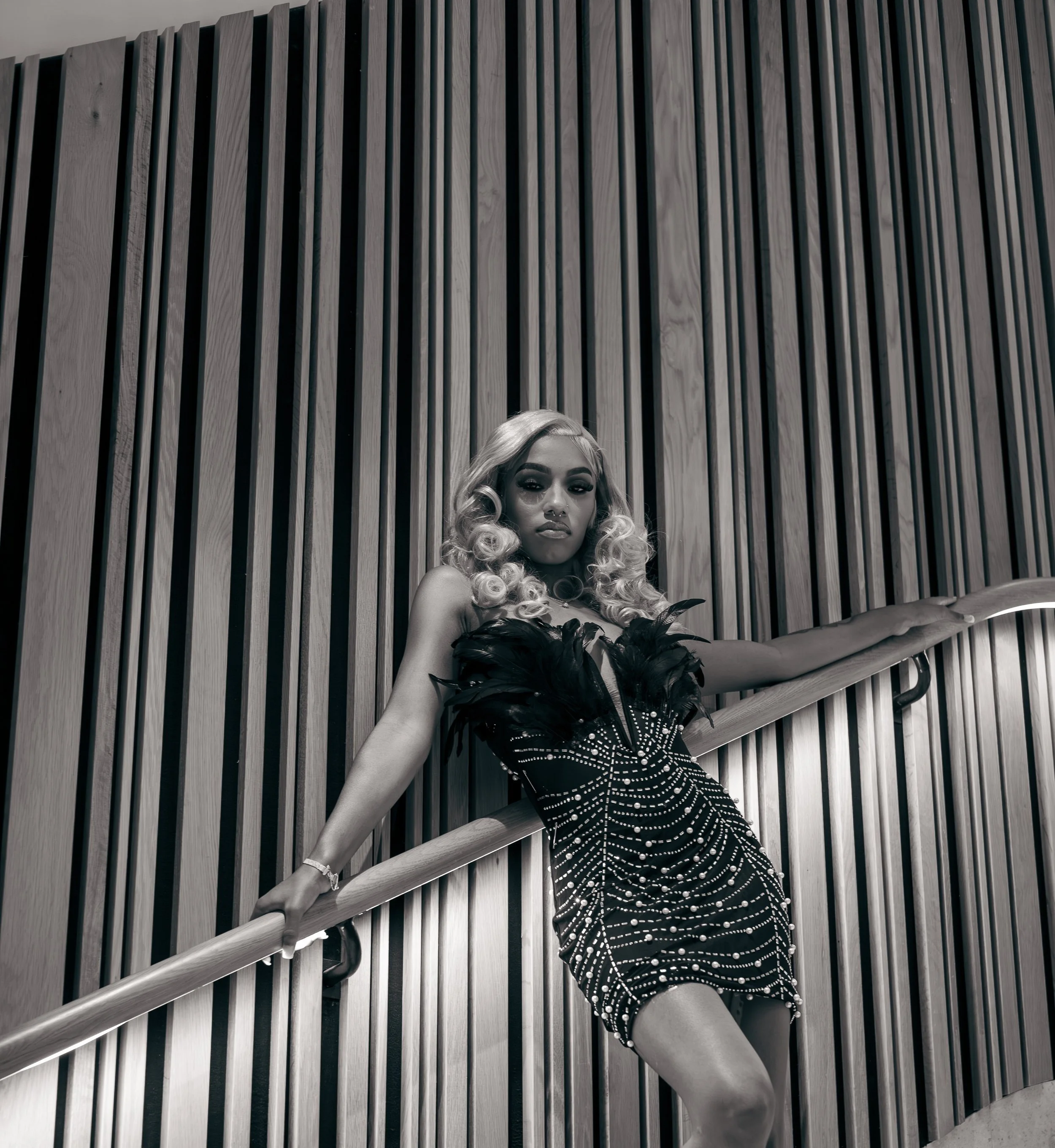 Person in a patterned dress with feathers, leaning against a railing, in front of a wooden slatted wall.