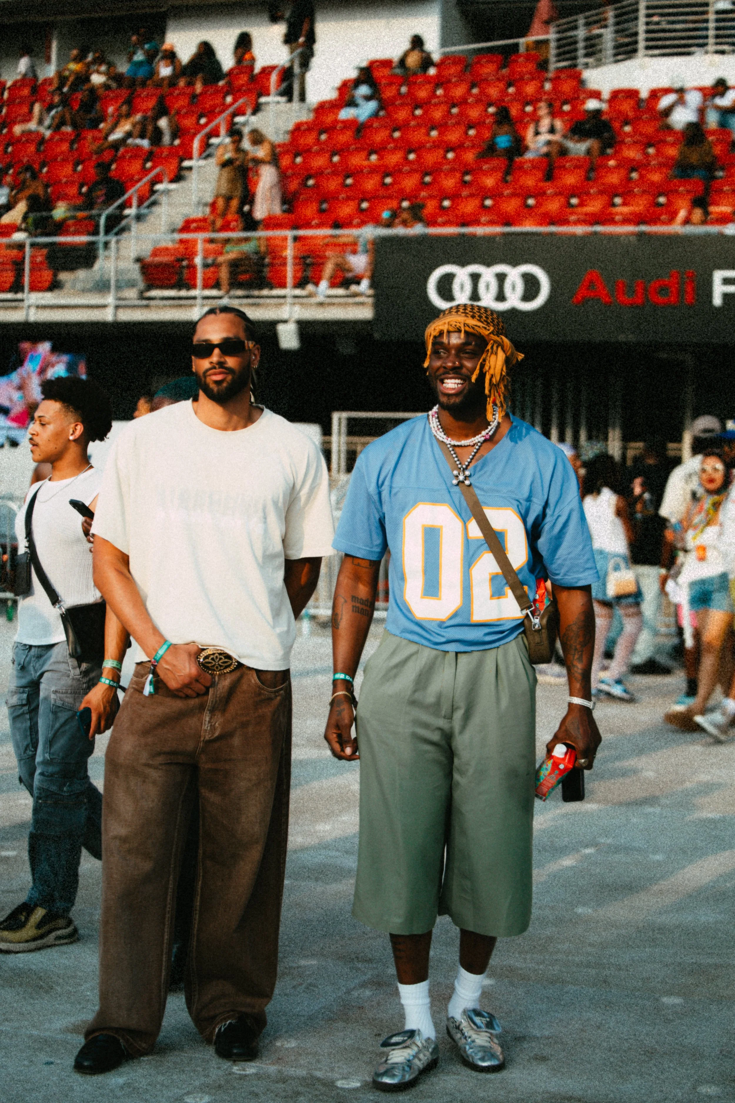 Two men standing in front of a stadium crowd; one in a white shirt and sunglasses, the other in a blue jersey and green shorts.