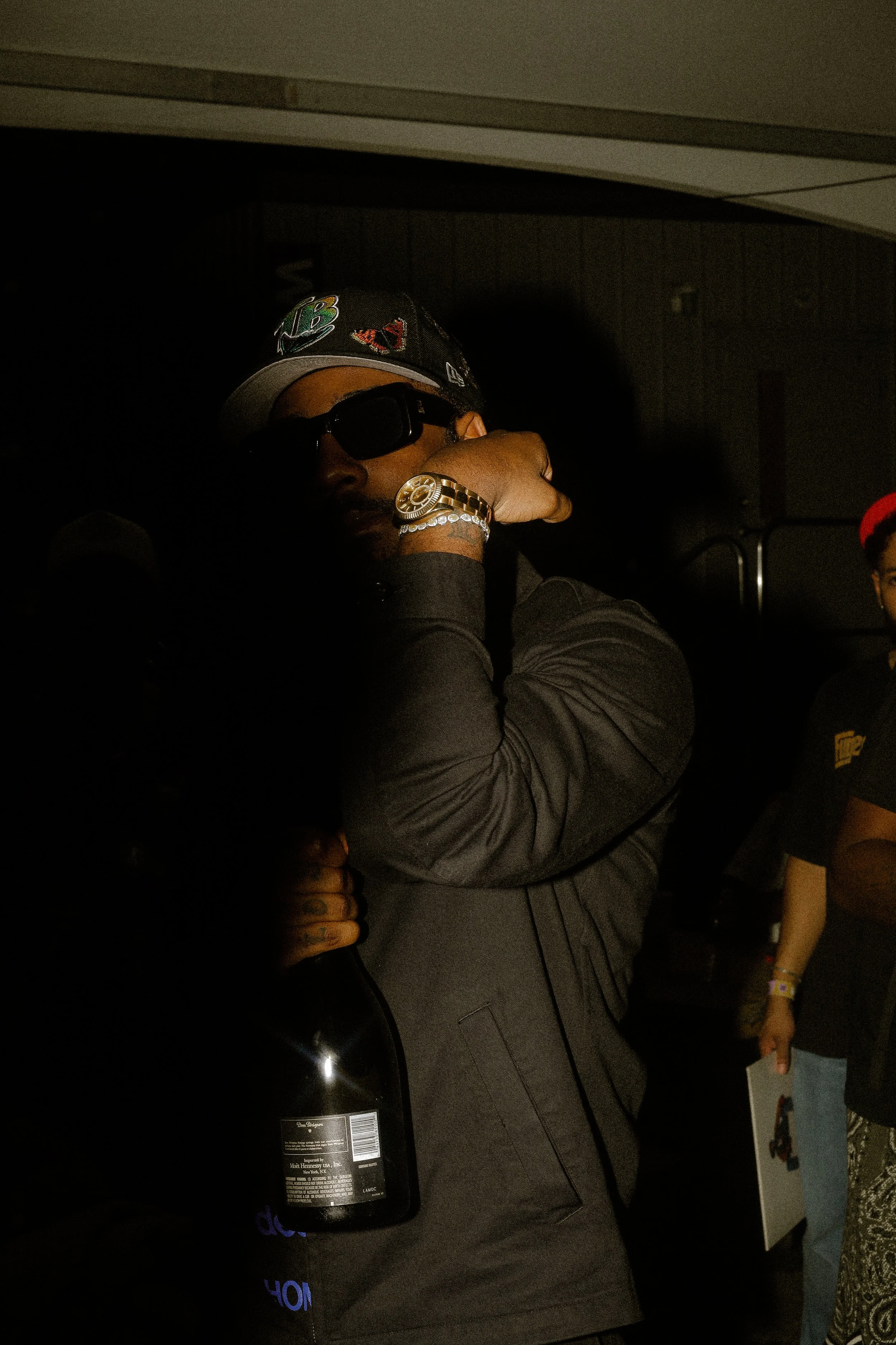 Person in dark clothing wearing sunglasses and a cap, holding a bottle, with a wristwatch visible.