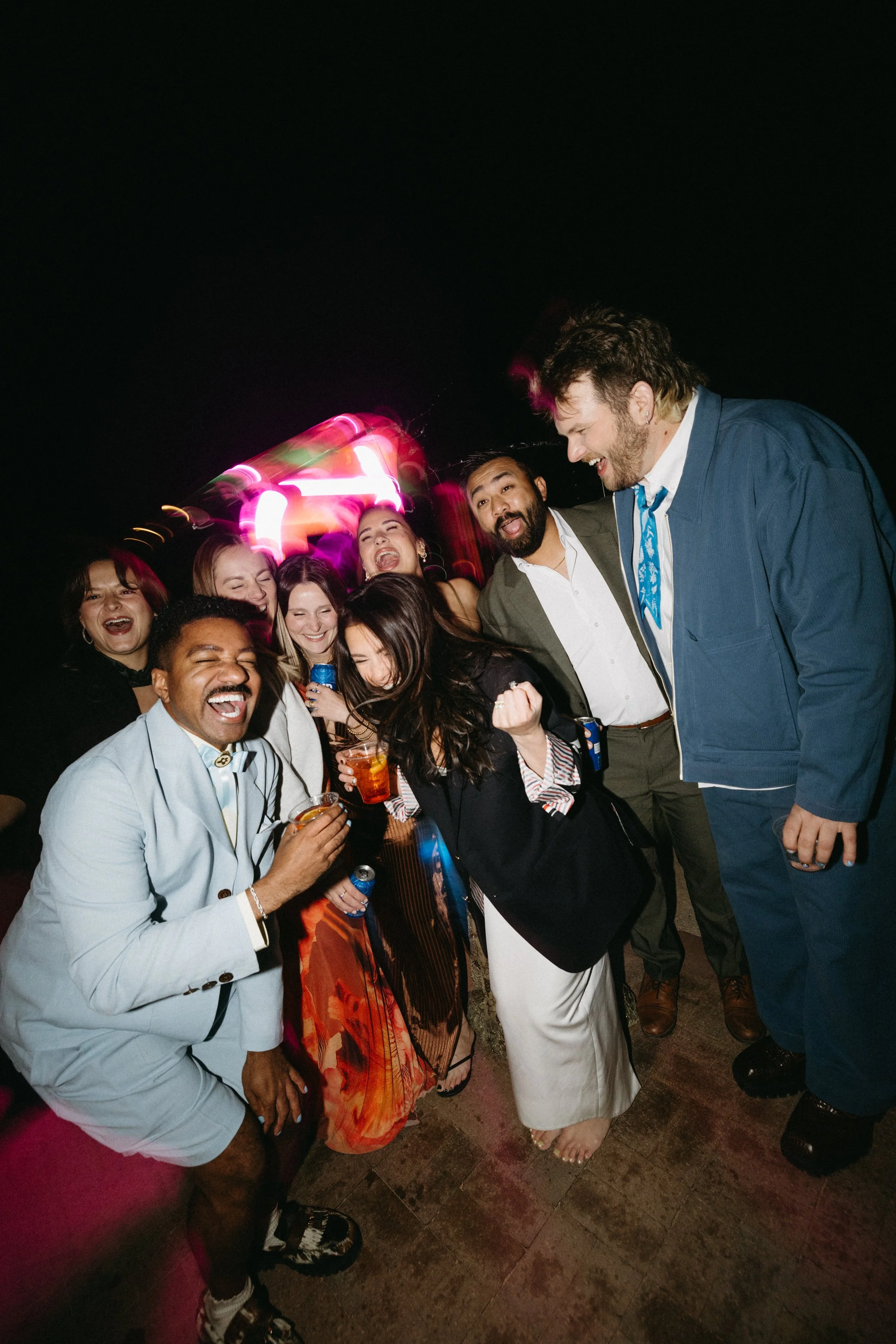 Group of diverse people celebrating at night, smiling, holding drinks, and dressed in fashionable clothing, with colorful lights in the background.