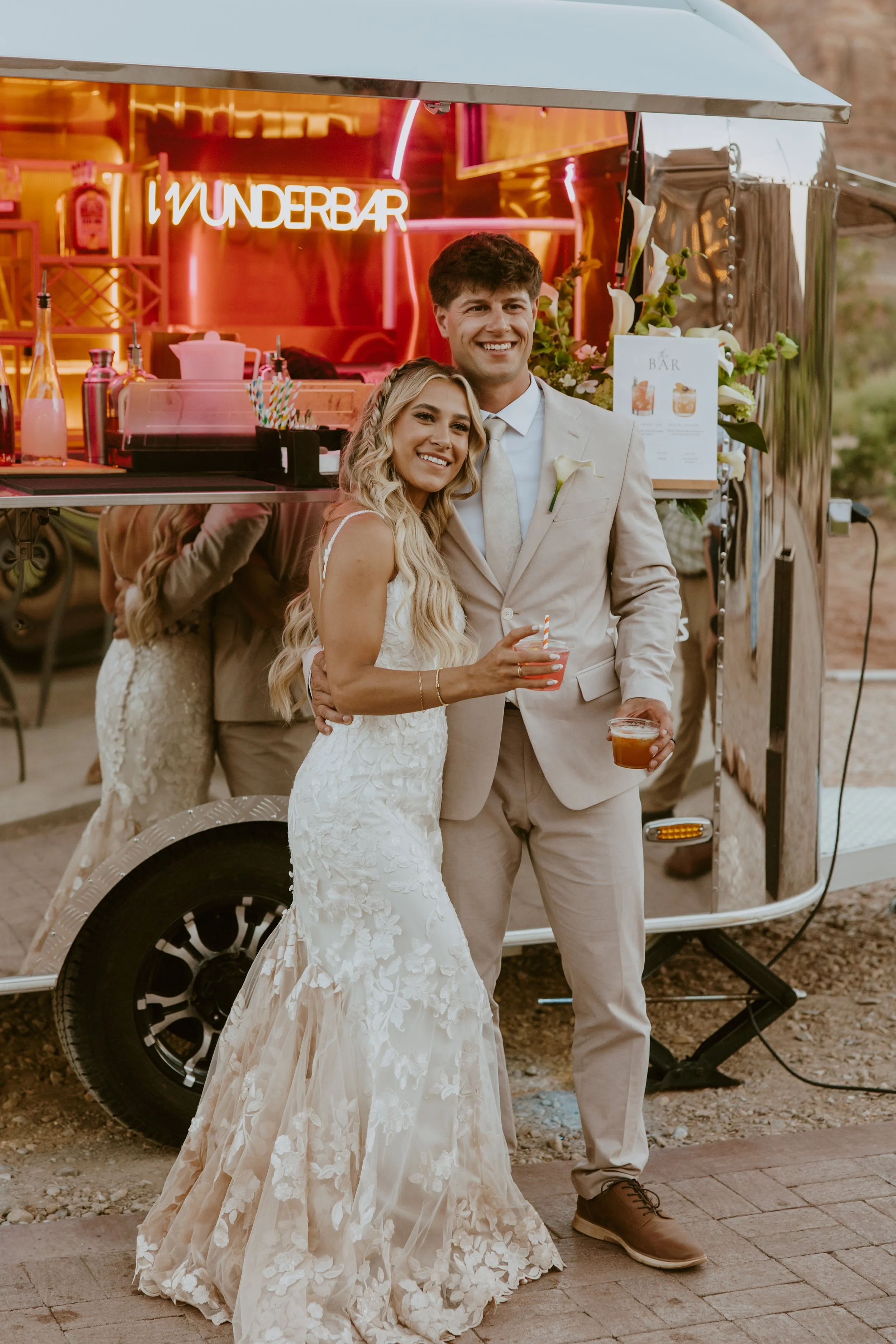 A smiling newlywed couple standing in front of a food truck. The bride is wearing a white lace wedding gown, and the groom is in a beige suit. They are holding drinks and posing for a photo, with the food truck behind them featuring a neon sign that 
