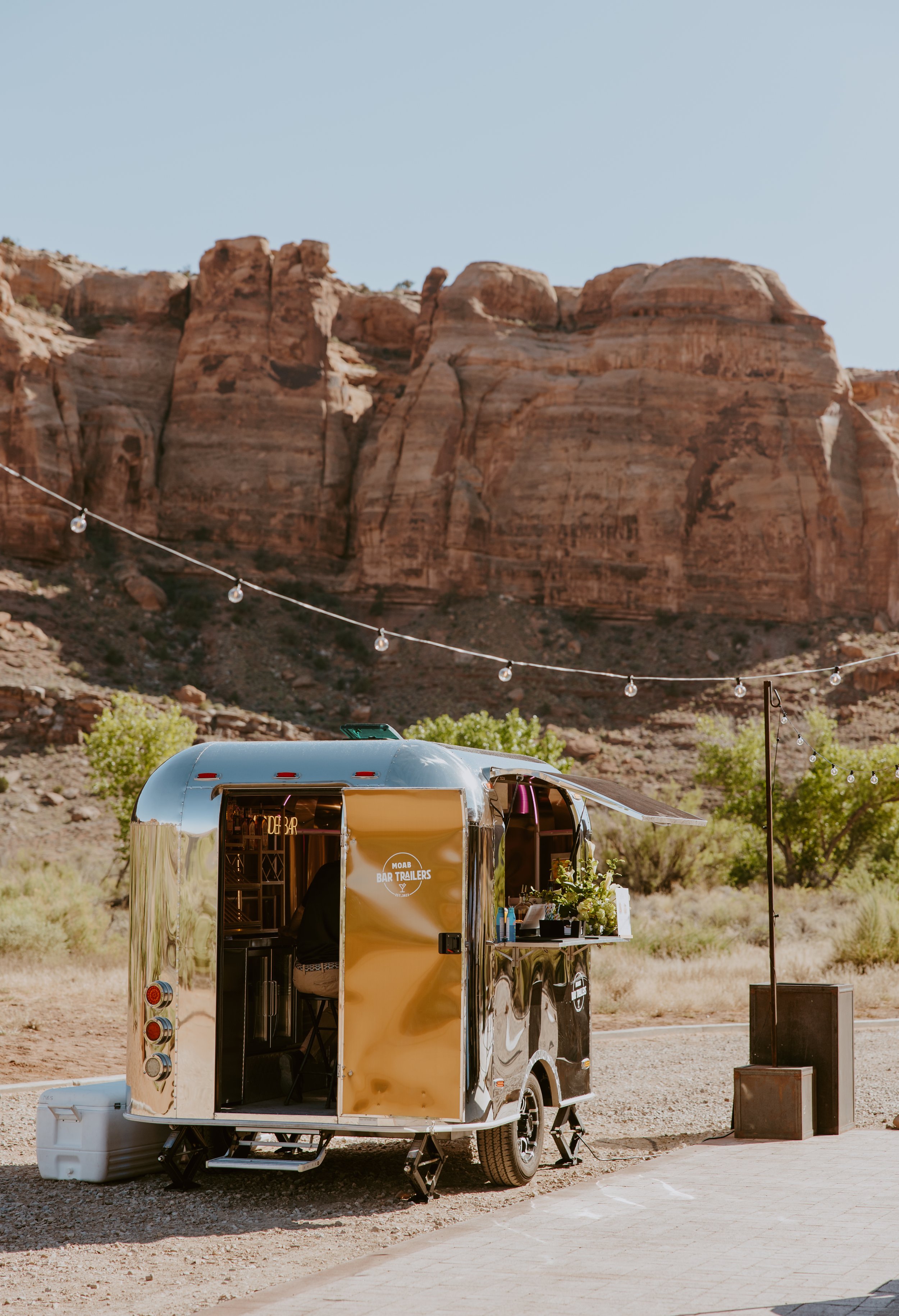 A shiny, metallic mobile bar trailer set up outdoors in a desert-like environment with large rock formations in the background. A string of lights is hung above the trailer, with a small table beside it and a food and drink sign on the side.