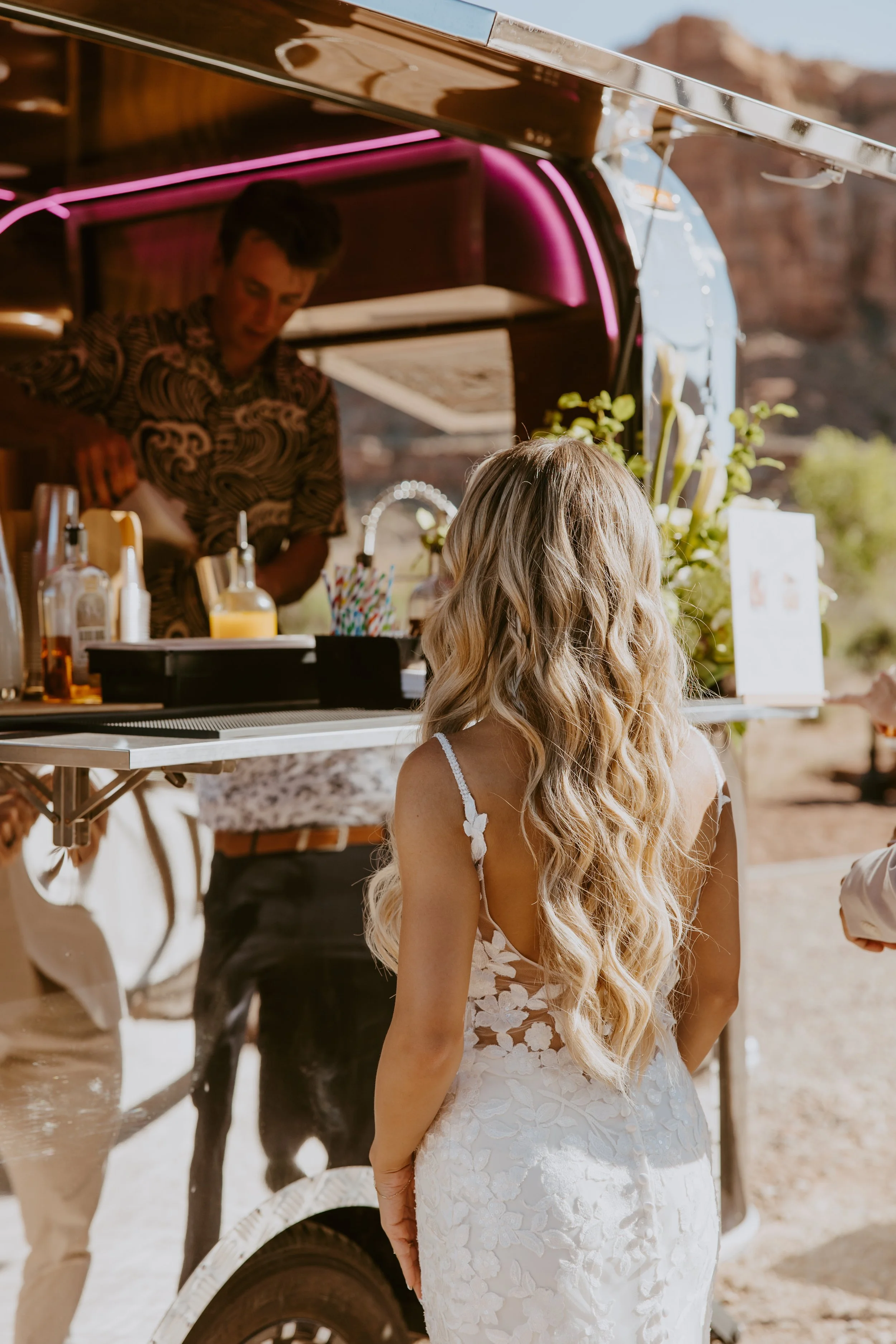 A woman in a white lace wedding dress with long blonde hair stands in front of a food truck at an outdoor event, with mountains in the background.