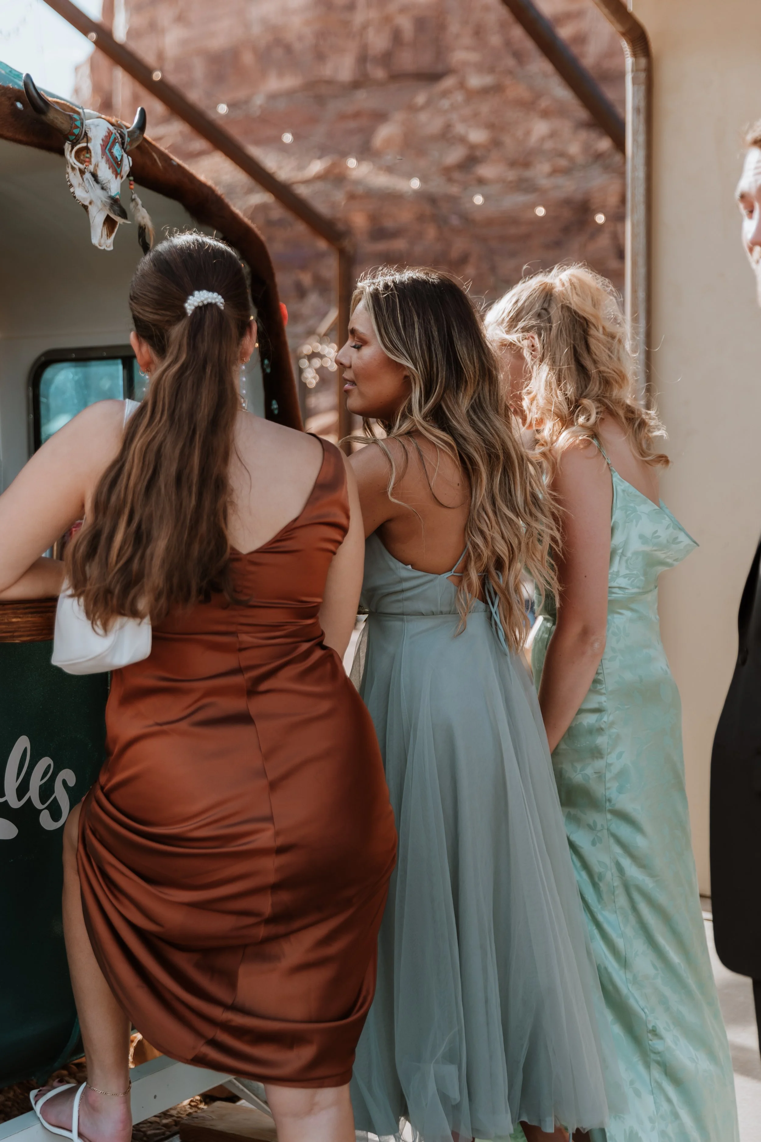 A group of women in colorful dresses standing close together, with their backs turned to the camera, near a food truck with a decorative animal skull with horns hanging on it, in a rustic outdoor setting.
