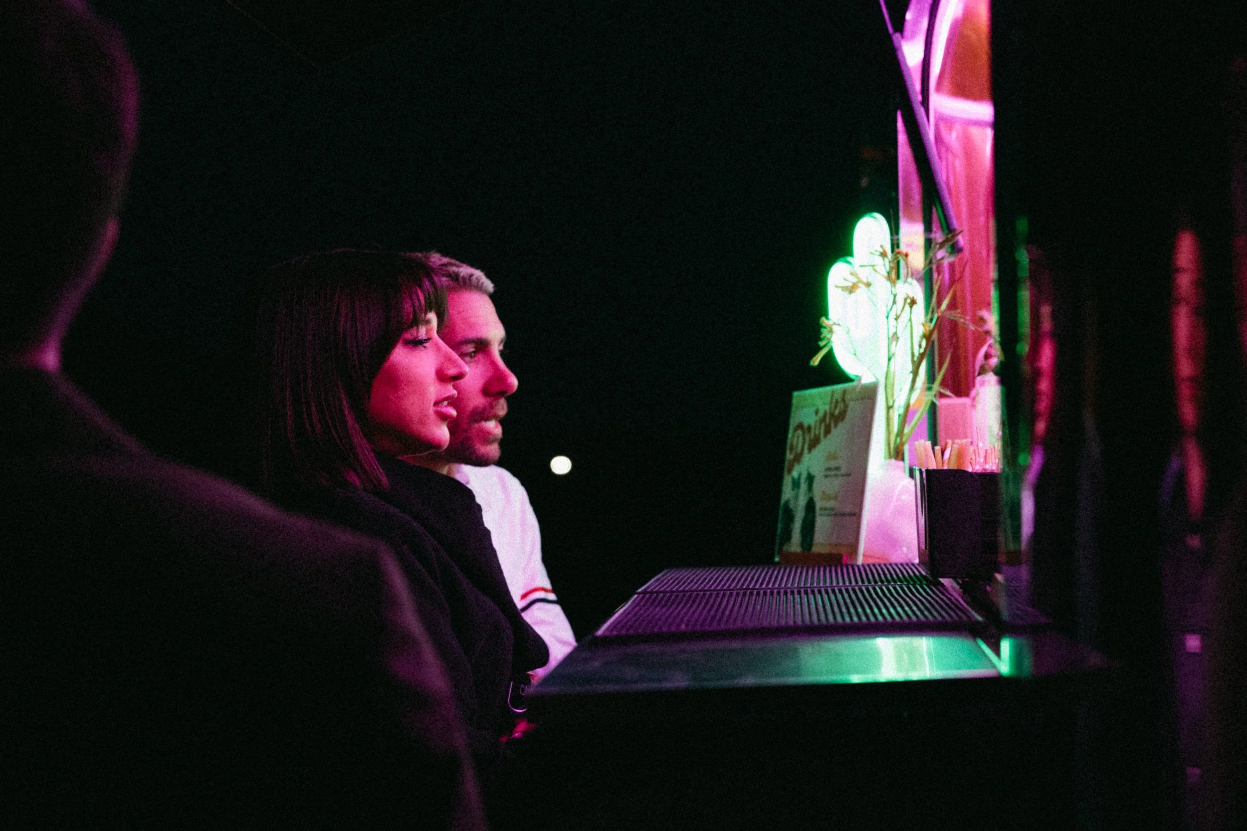 Profile of three people sitting at a neon-lit bar or counter at night, with colorful neon signs and decorative plants in the background.