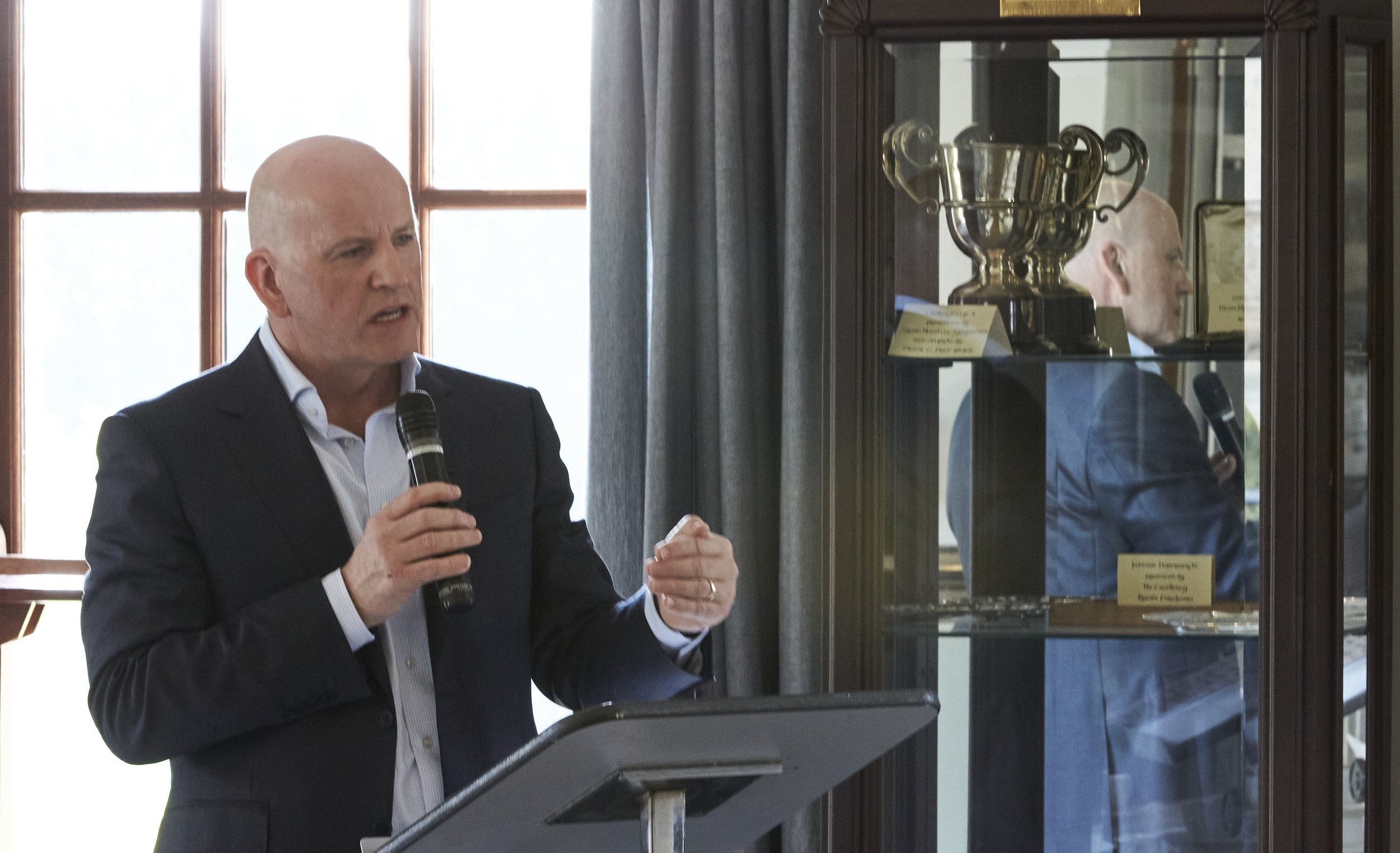 Man in suit speaking into a microphone at a podium, with a trophy displayed in a glass cabinet behind him.