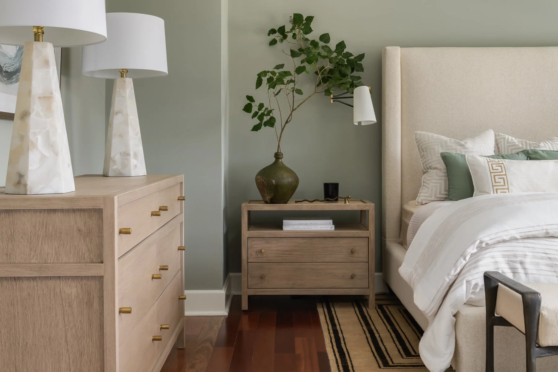A bedroom corner with a beige upholstered bed, a wooden nightstand with a large green vase and a plant, and a dresser with two white lamps on top.