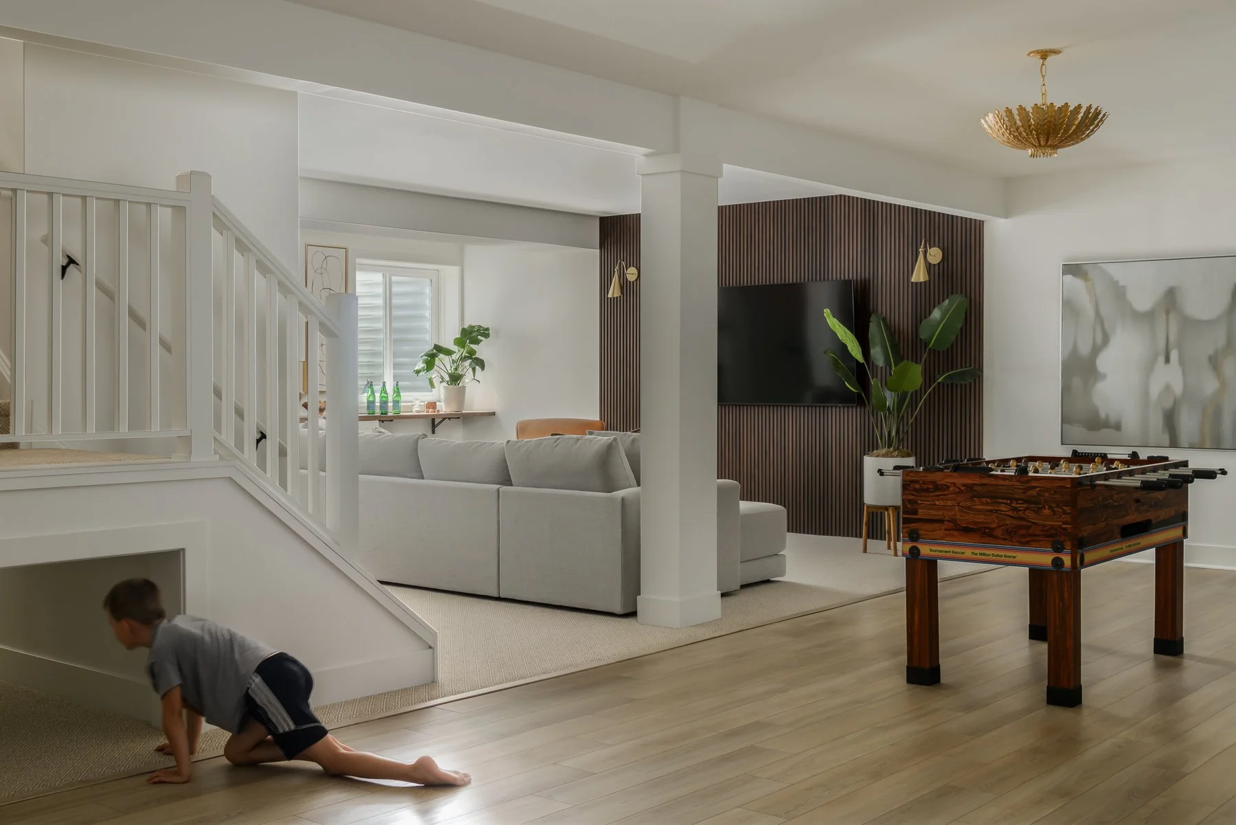 A living room with white walls, a beige area rug, a light gray sectional sofa, a wooden coffee table with foosball game, a large flat-screen TV mounted on a wood-paneled accent wall, tropical plant, framed art, and window with blinds. There is a chil