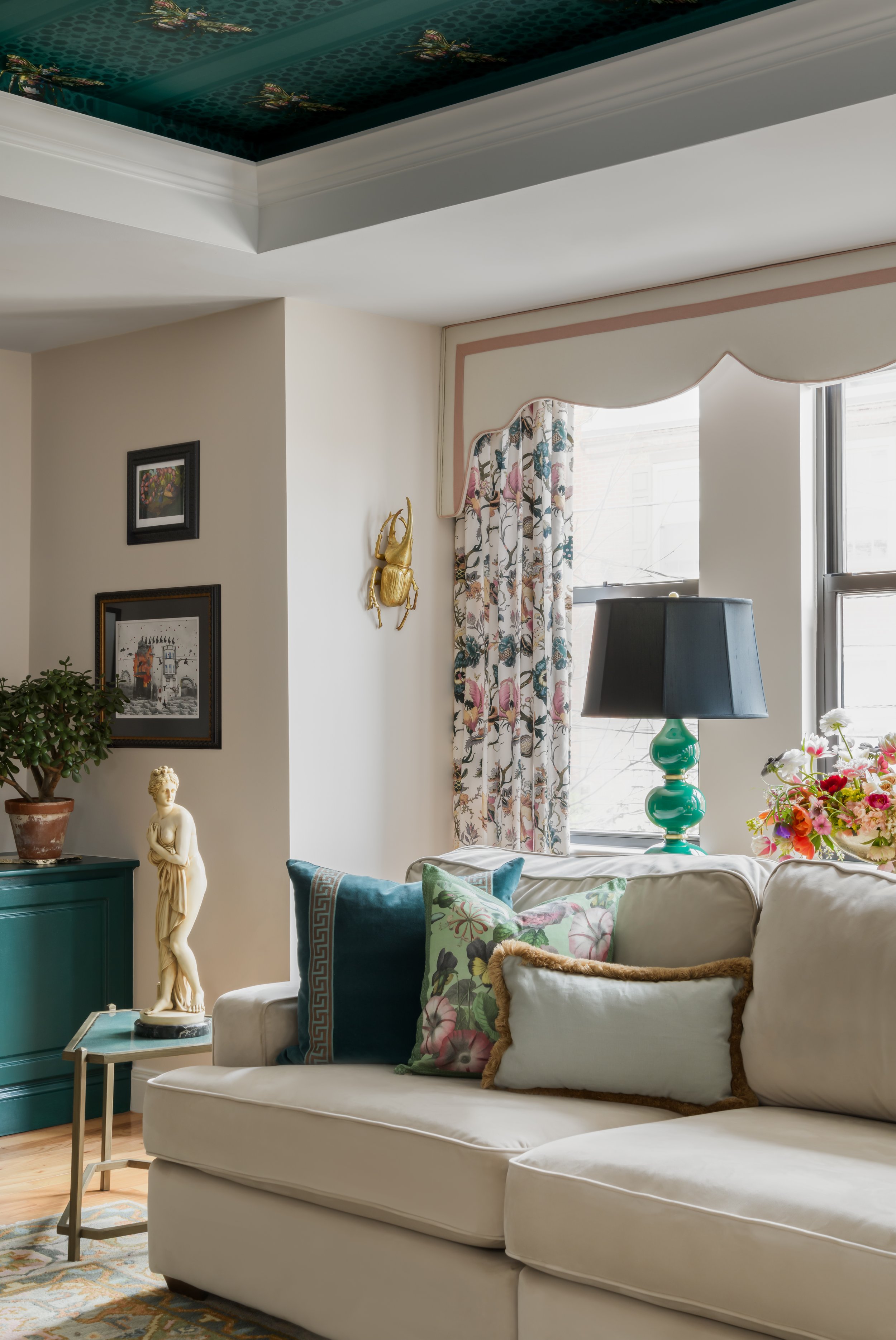 A living room with a white sofa, decorative pillows, a green lamp, floral curtains, a small table with a statue, framed art on the wall, and a large window.