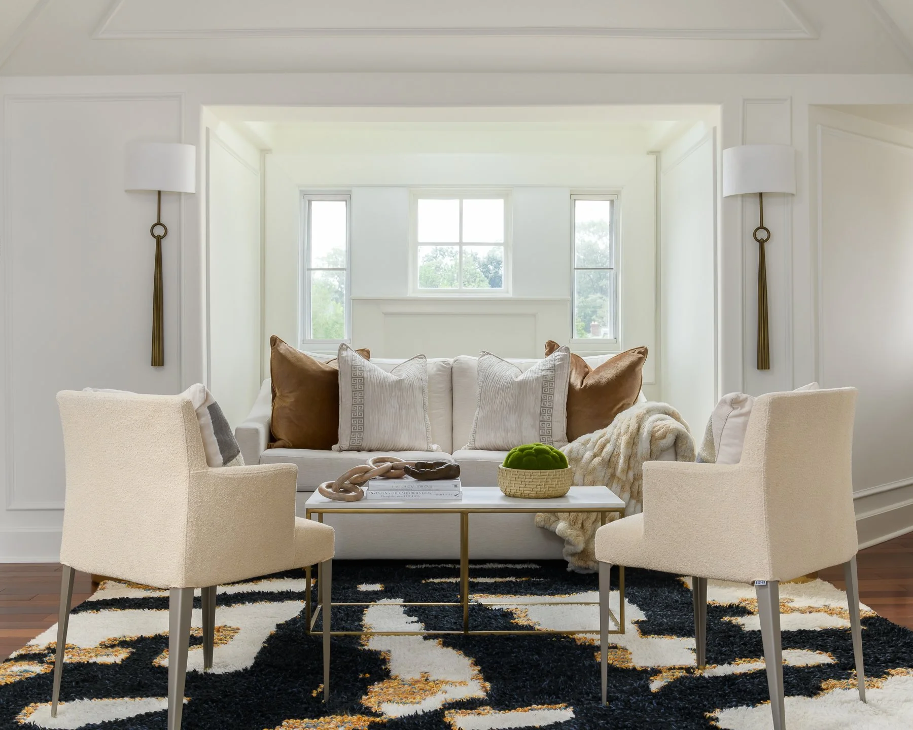 A bright living room with white walls and large window. Centered is a white sofa with beige and brown cushions. In front of the sofa, a white and gold coffee table holds books, decorative objects, and a green moss centerpiece. Two beige armchairs sur