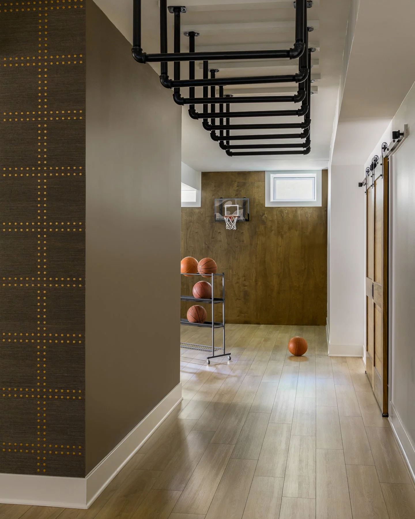 Indoor basketball court with basketball hoop on the back wall, basketballs on a cart, and a ball on the wooden floor, with black pipe storage structures hanging from the ceiling.