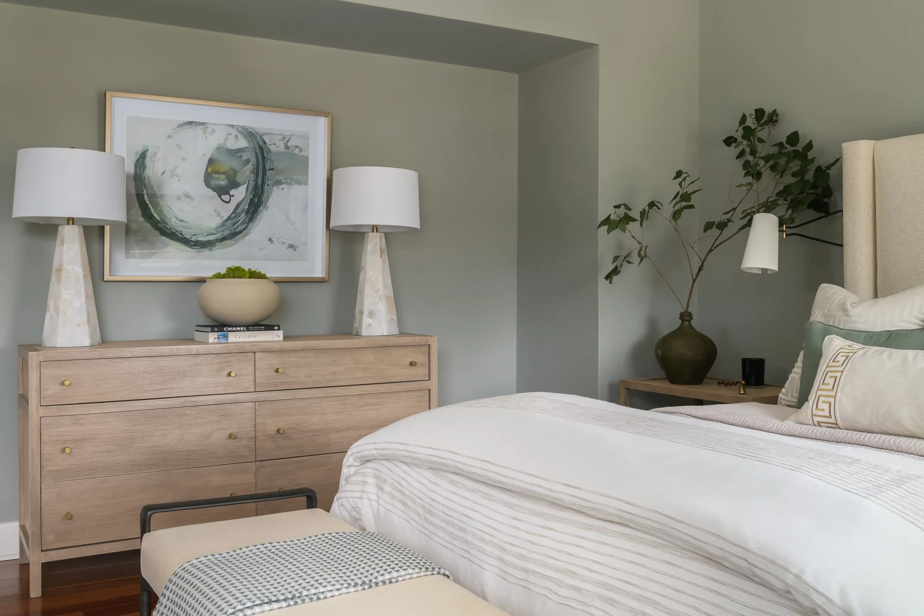 A tidy bedroom with a light wood dresser topped with two white lamps, a framed abstract painting, and a beige bowl with greenery. A bed with white linens, decorative pillows, and a headboard with a wall-mounted reading lamp is visible. A nightstand w
