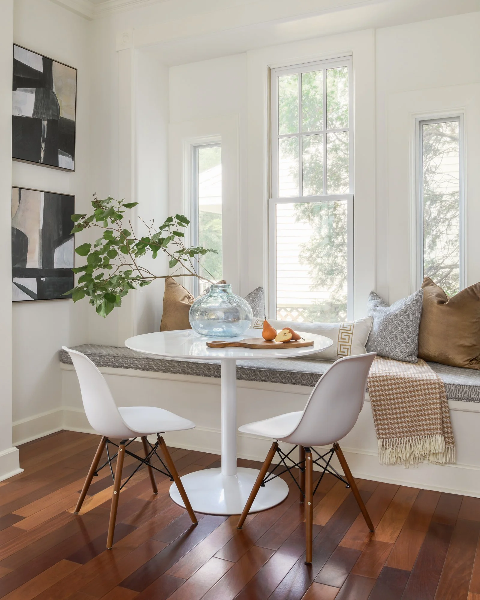 Sunlit dining nook with a white round table, two modern white chairs, a window bench with assorted pillows, artwork on the wall, and hardwood flooring.