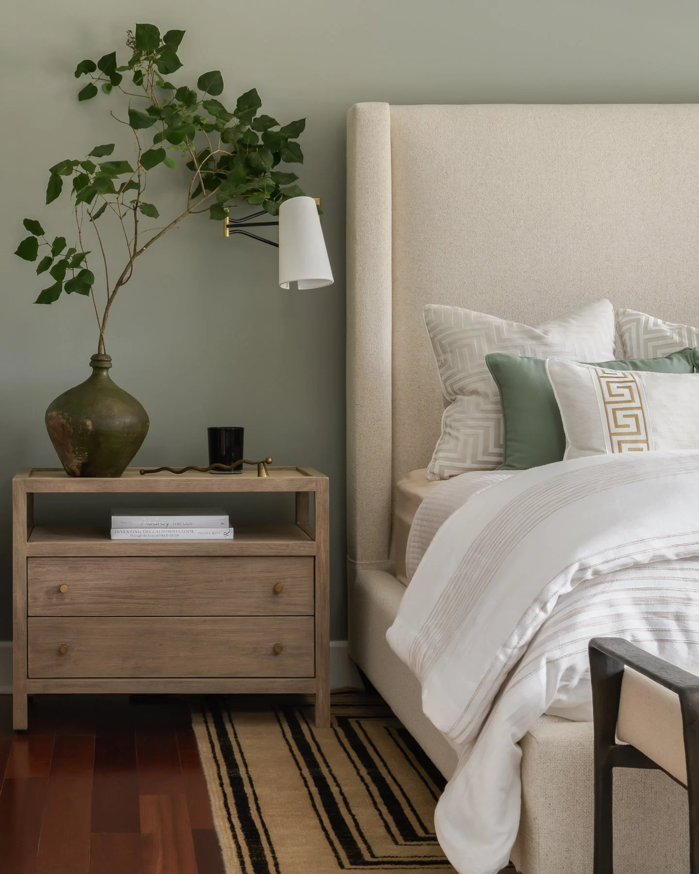 A bedroom scene with a beige upholstered bed, white and patterned pillows, a wooden nightstand with a large vase of green leaves, a black cup, and a small lamp. The room has a neutral color palette with soft green walls and a black and beige striped 