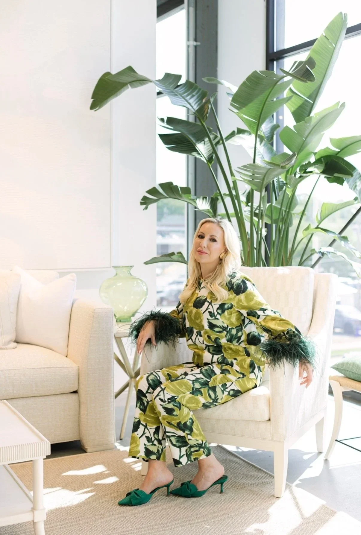 A woman in a yellow and green floral outfit sitting on a white couch in a bright room with large windows and green plants.
