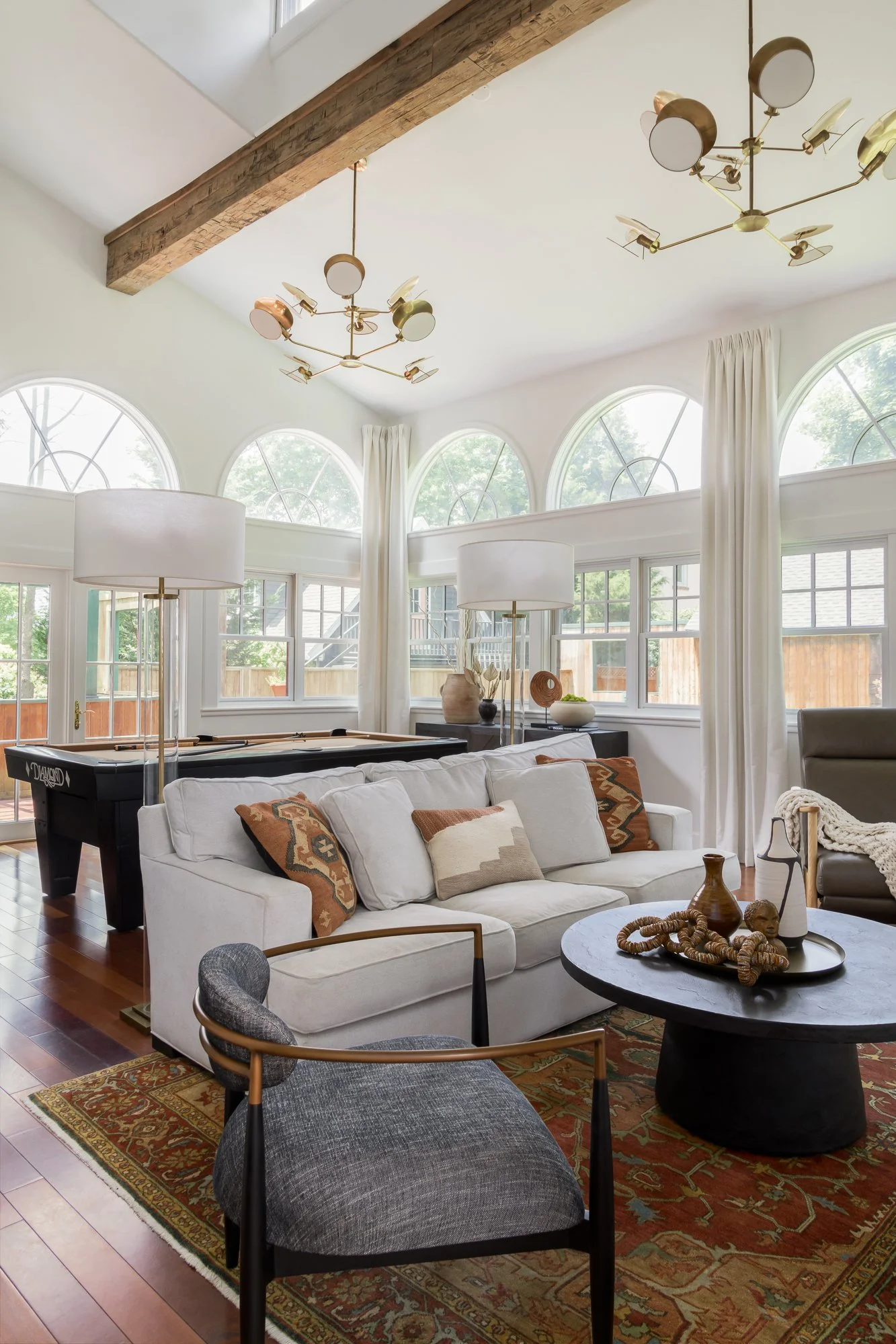 Living room with white sofa, patterned pillows, black coffee table with decorative objects, arched windows, cream curtains, leather armchair, floor lamps, artistic ceiling lighting, hardwood floors, area rug, and large wooden beam ceiling detail.