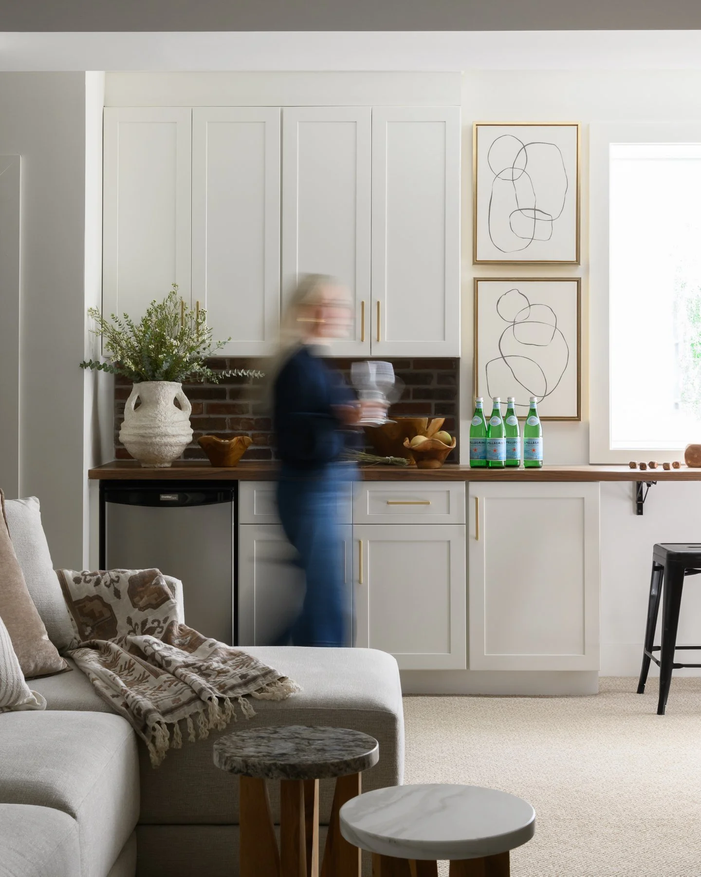 Living room with white sofa, beige and brown pillows, and small tables. In the background, a blurred person in dark clothing holds a glass in front of a kitchen counter with decorative items, plants, and framed artwork on the wall.