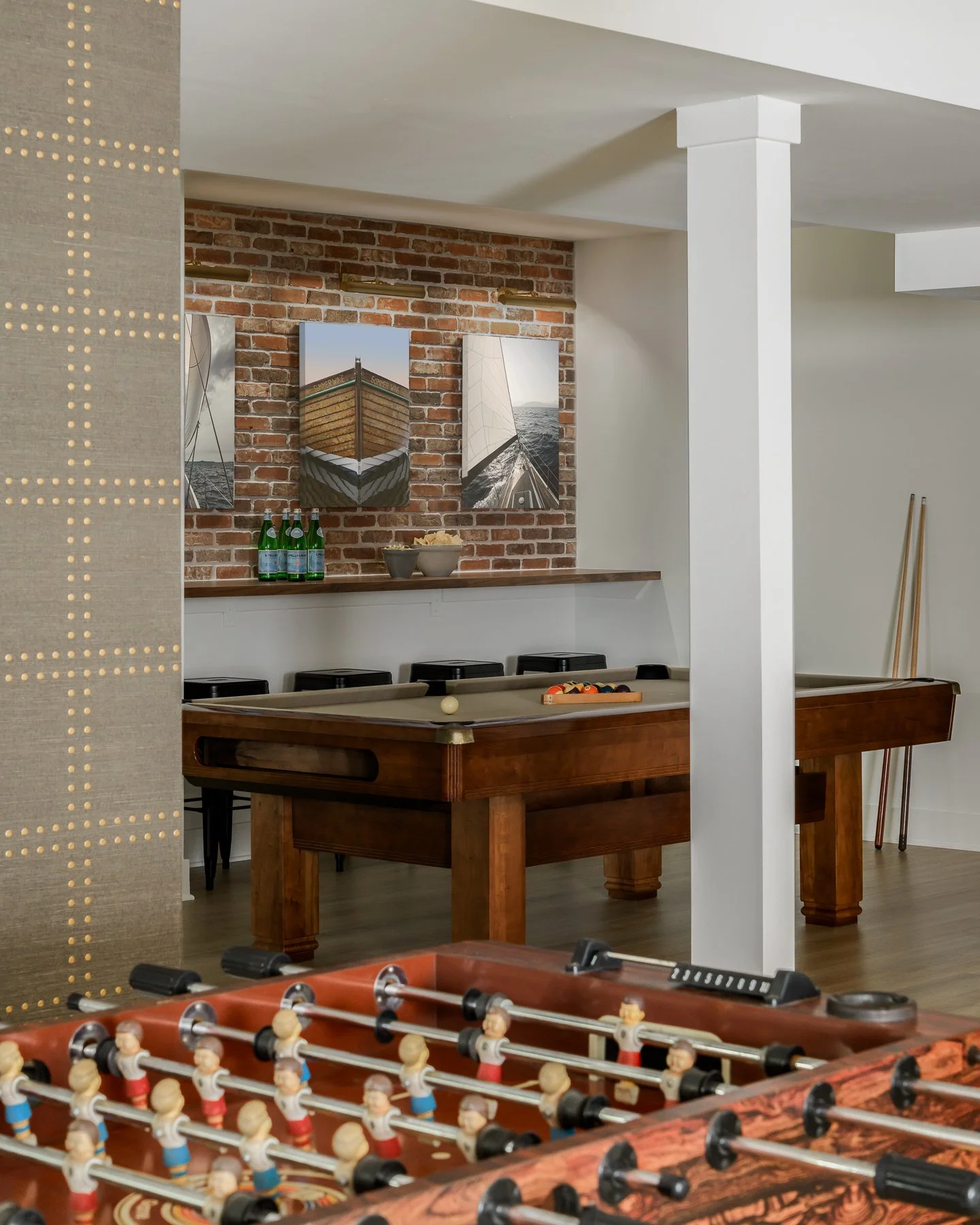 A room with a brick accent wall, decorated with three framed nautical-themed photographs, a pool table with a set of pool balls, and four bottles of sparkling water on a wooden shelf.
