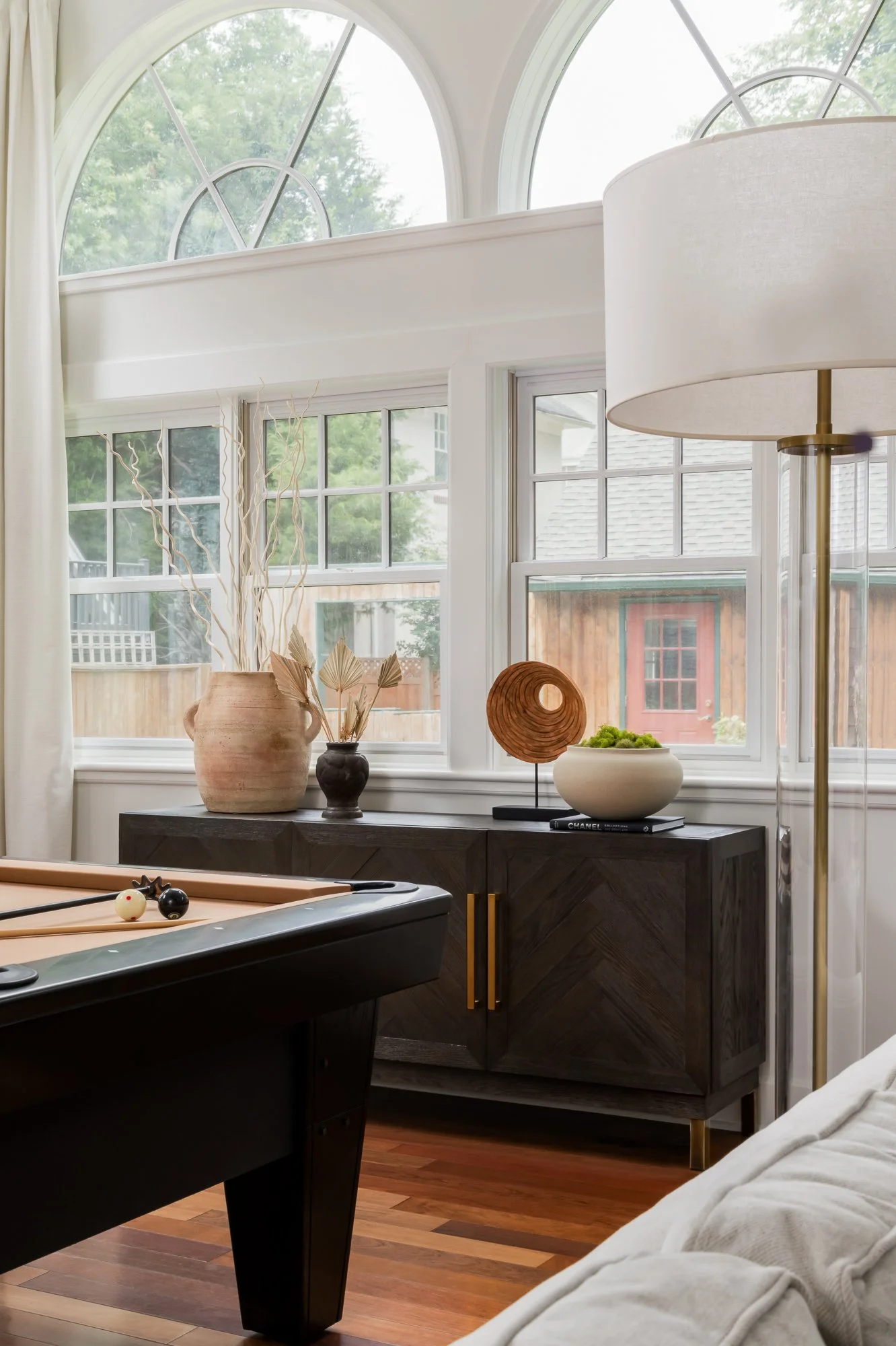 Living room with large arched windows, decorative vases on a dark wood sideboard, a pool table with cue balls, a floor lamp, and a white sofa.