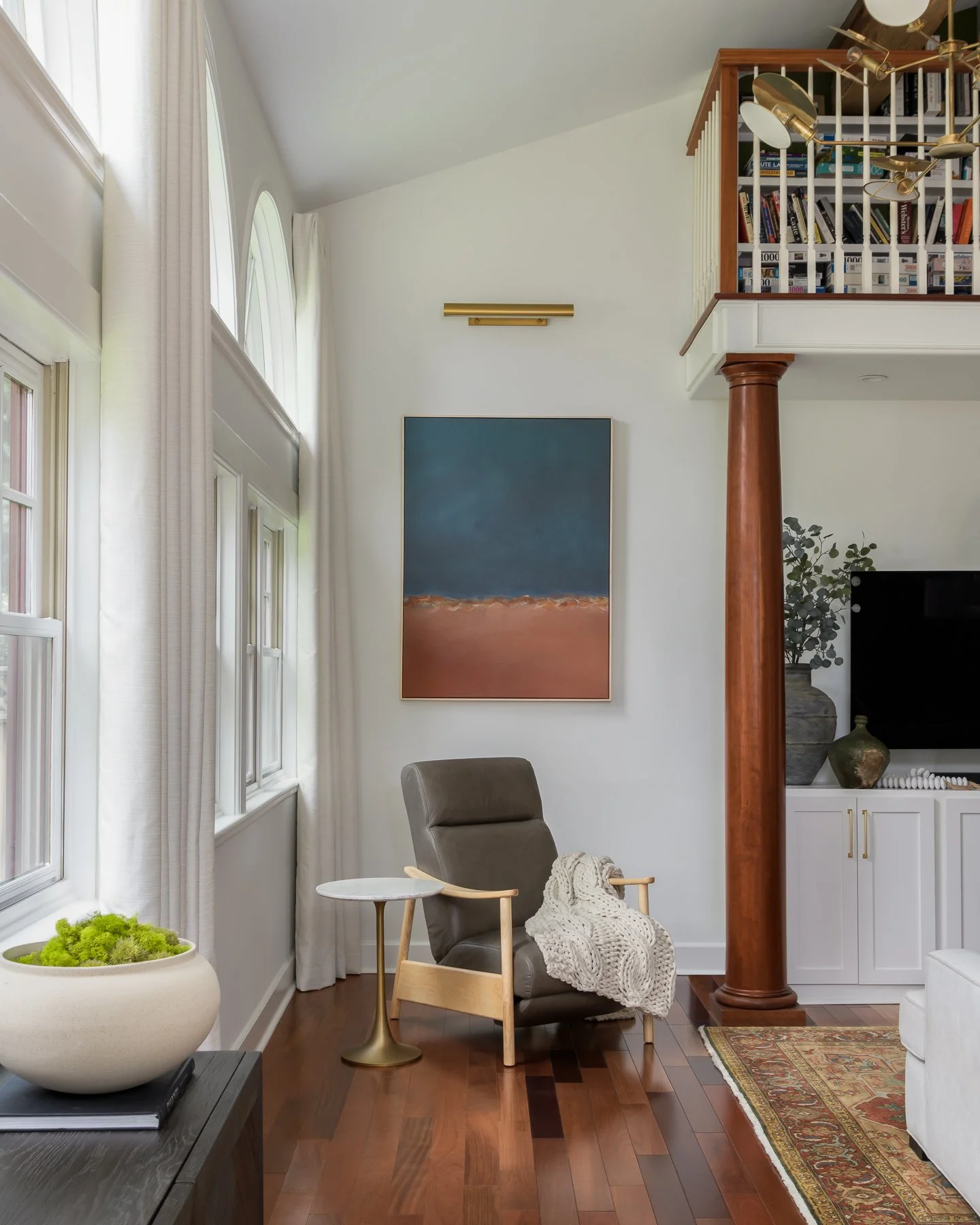 Interior view of a living room with large windows, white curtains, a beige armchair with a knit blanket, a small round side table, a painting on the wall, bookshelf, and decorative plants.