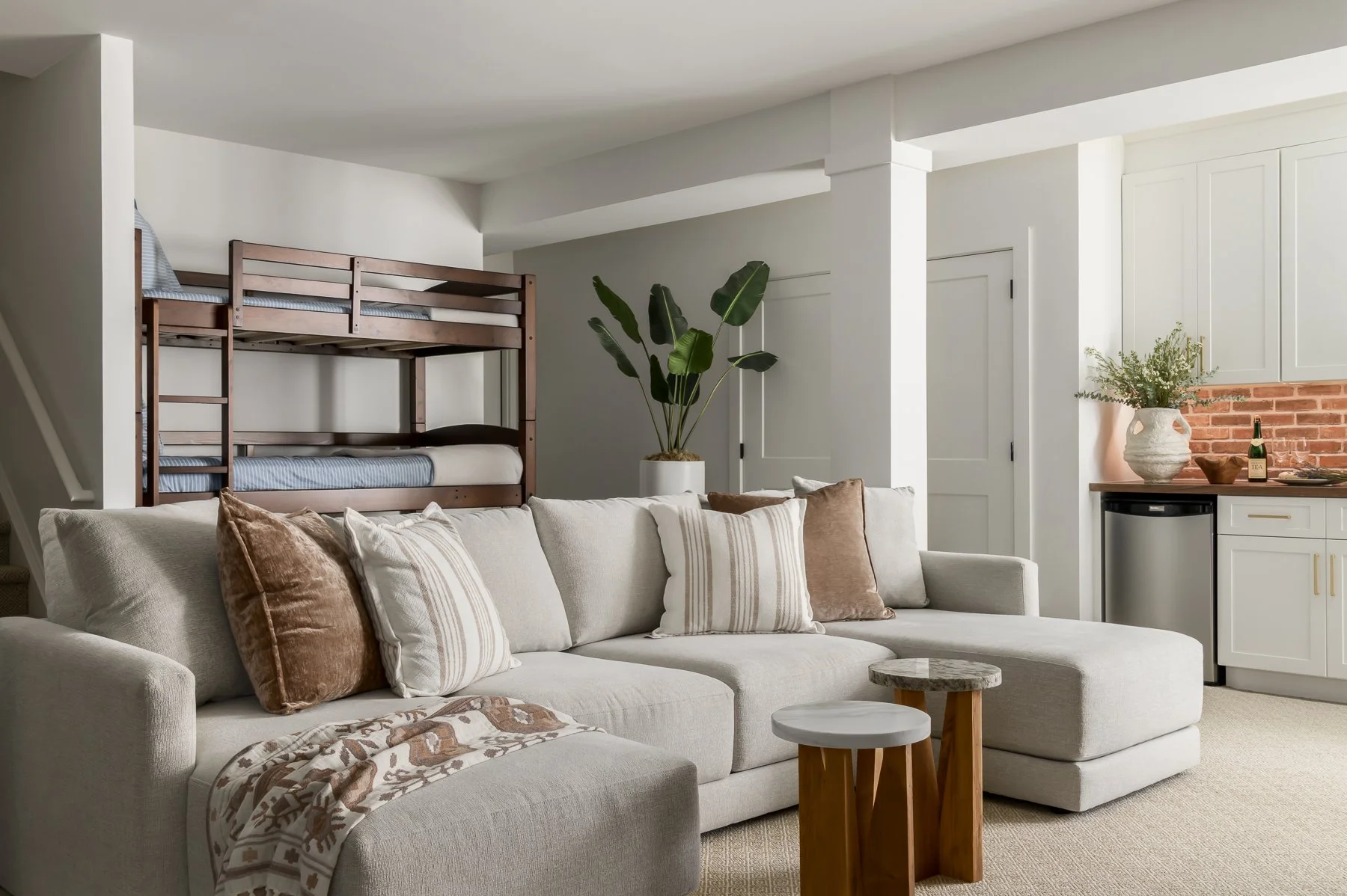 Living room with cream-colored sofa decorated with brown and striped pillows, a small wooden side table, a large houseplant in a white pot, and a kitchenette with white cabinets, a small mini fridge, a potted plant, and a tray with a bottle and a bow
