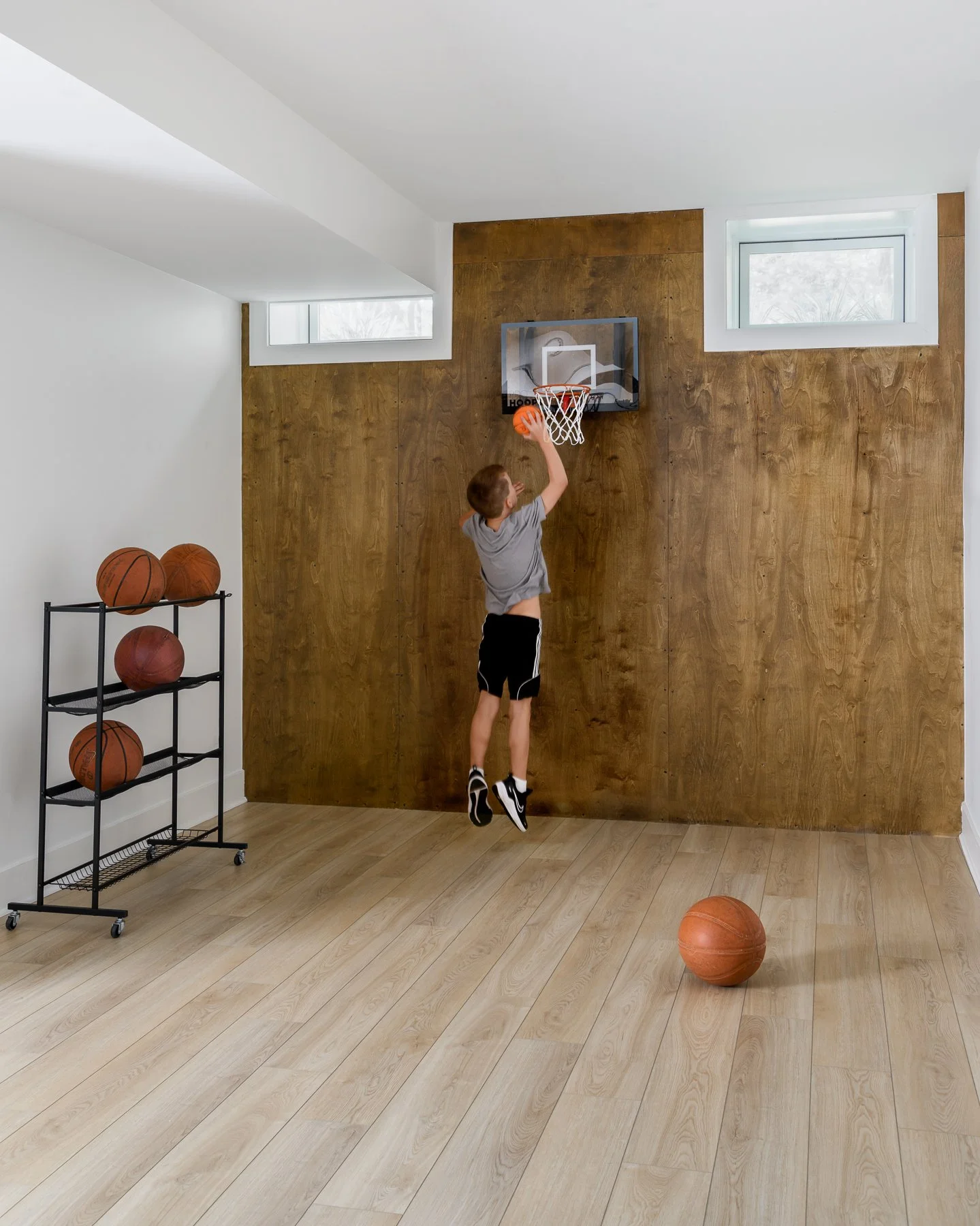 A boy in a gray shirt and black shorts practicing basketball in a home gym, jumping towards a hoop mounted on a wooden wall, with basketballs on a rack and floor around him, and two small windows in the background.