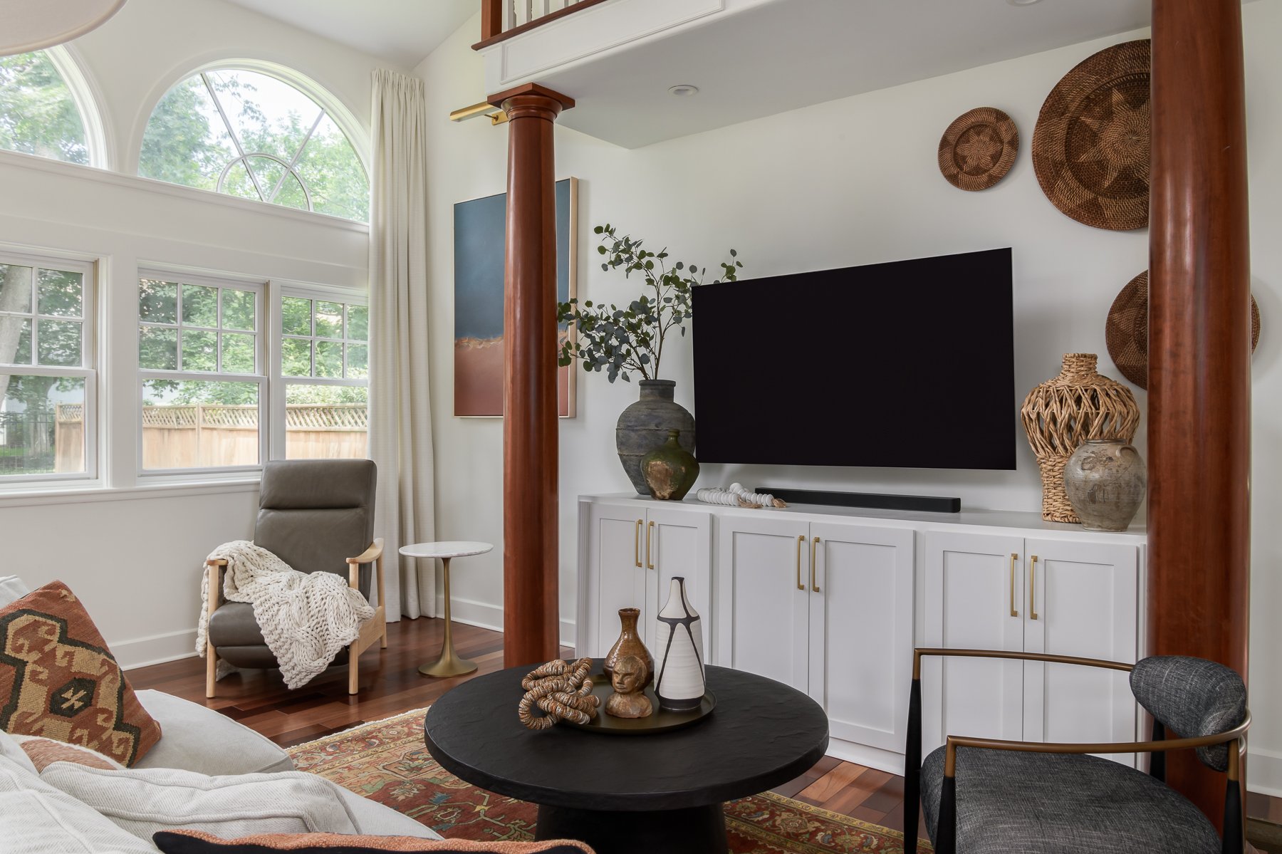 Living room with large windows, a white cabinet with decorative vases, a flat-screen TV, a gray armchair with a blanket and pillow, and various decorative baskets and artwork on the wall.