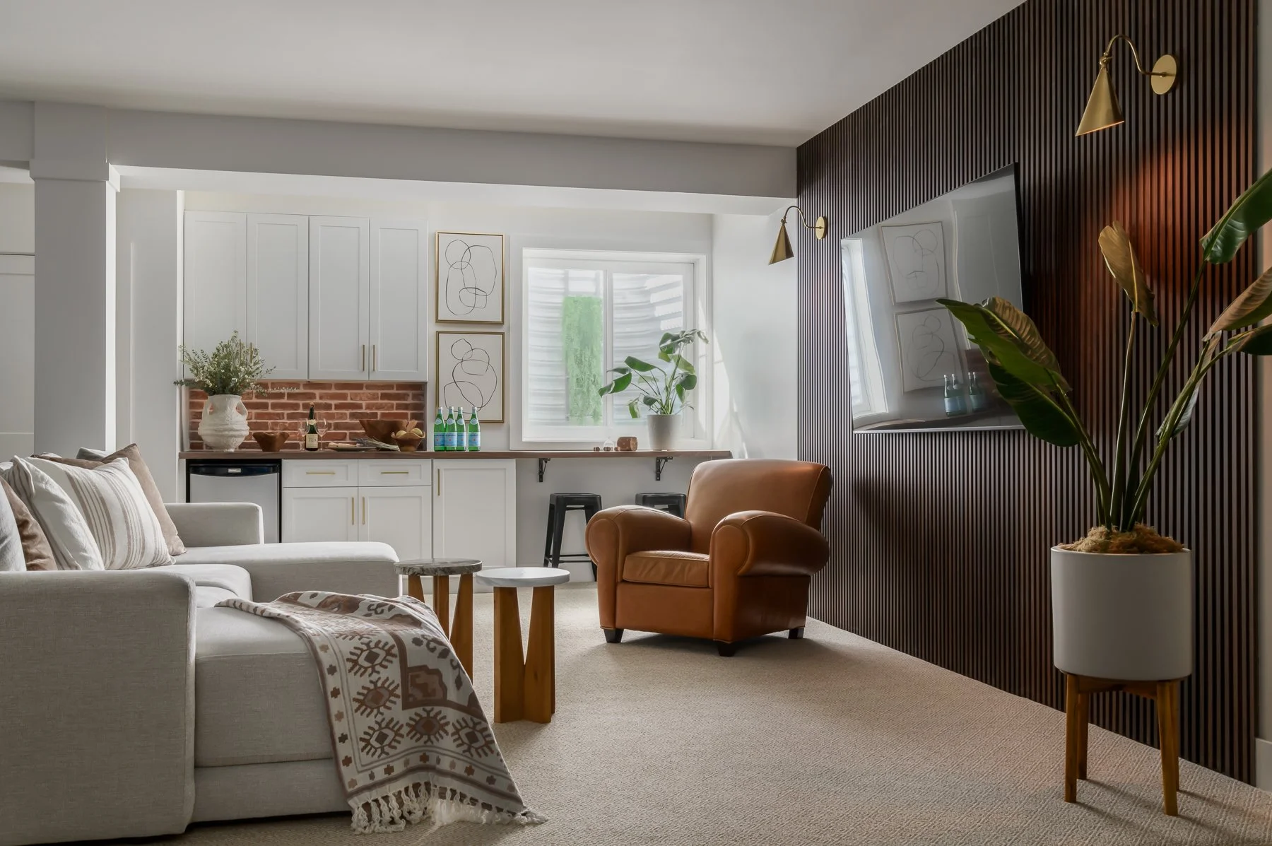 Living room with white sofa, brown armchair, wooden side tables, and a large plaid blanket. Modern wall art, potted plants, and a window with sunlight. Kitchen area in the background with white cabinets, a brick backsplash, and bar stools.