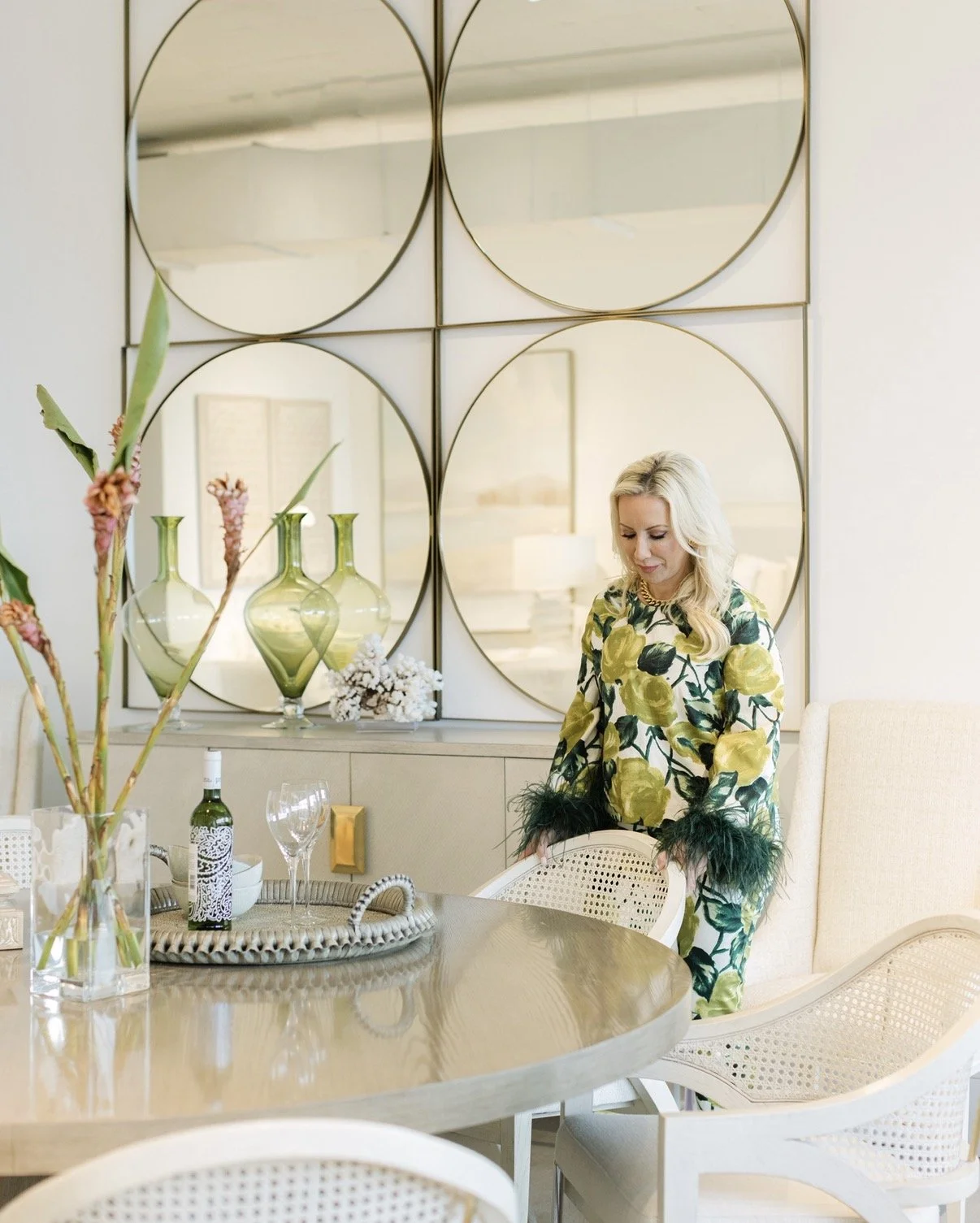 A woman in a floral yellow and green dress with black feathered cuffs standing by a dining table in a modern, elegant room with wall mirrors, vases, and decorative objects.