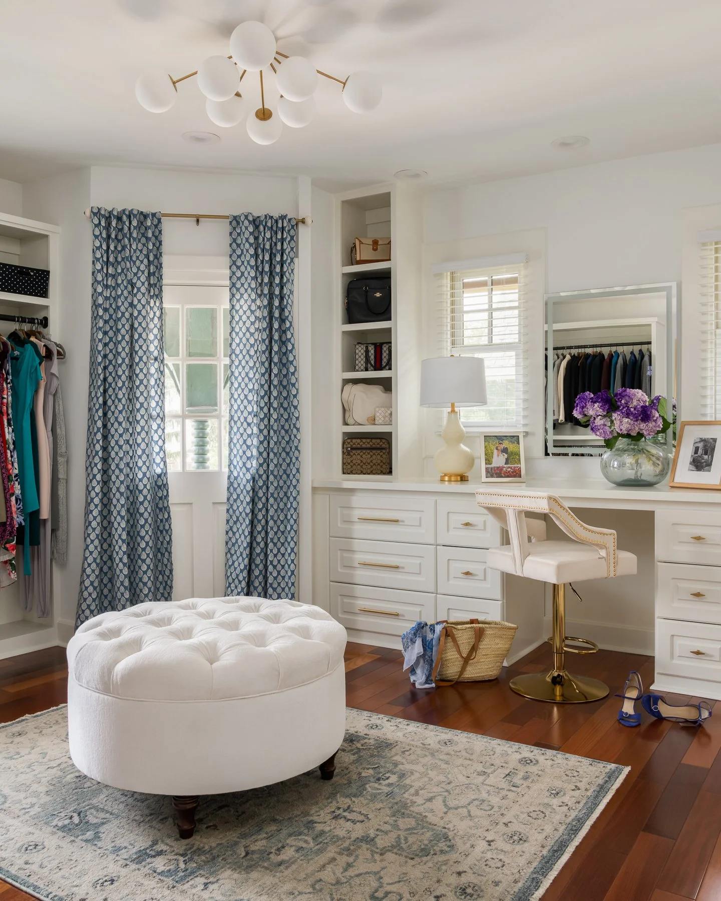 A bright and stylish walk-in closet with white cabinetry, a marble top vanity, a cushioned white ottoman, and hardwood floors. Blue patterned curtains hang over a window, and there are various bags on shelves and a framed photo on the vanity with a b