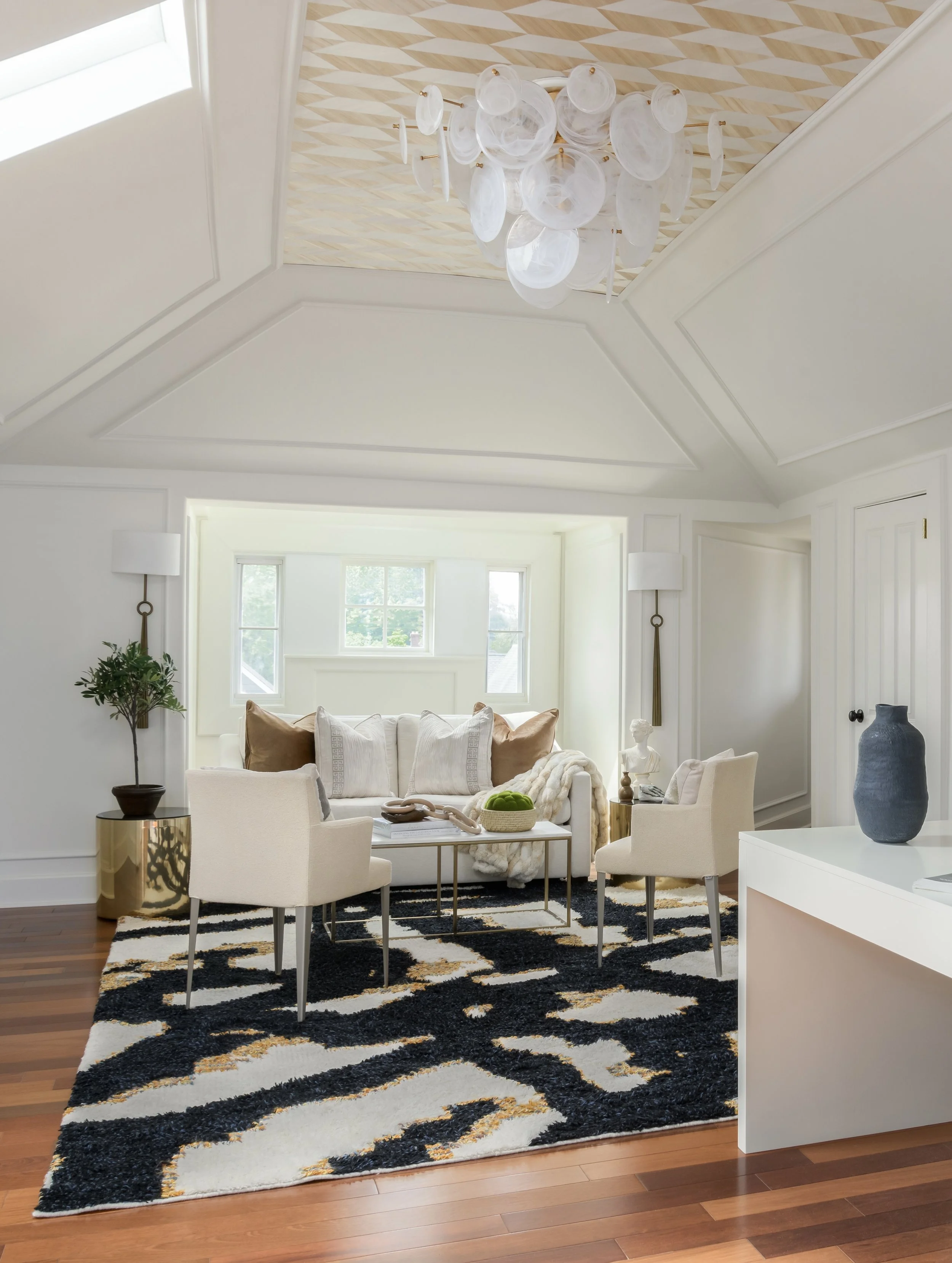 A living room with a high, white ceiling with molding, a black and white striped rug on a hardwood floor, and a sitting area with a white sofa, beige armchairs, and a glass coffee table. The room is decorated with a large abstract white chandelier, a