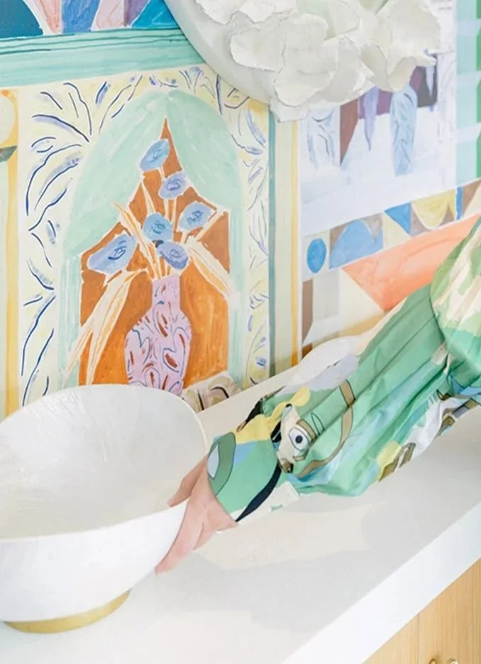 Child reaching into a white bowl with a colorful long sleeve shirt, standing in front of a painted mural depicting a bouquet of blue flowers in an orange and pink vase.