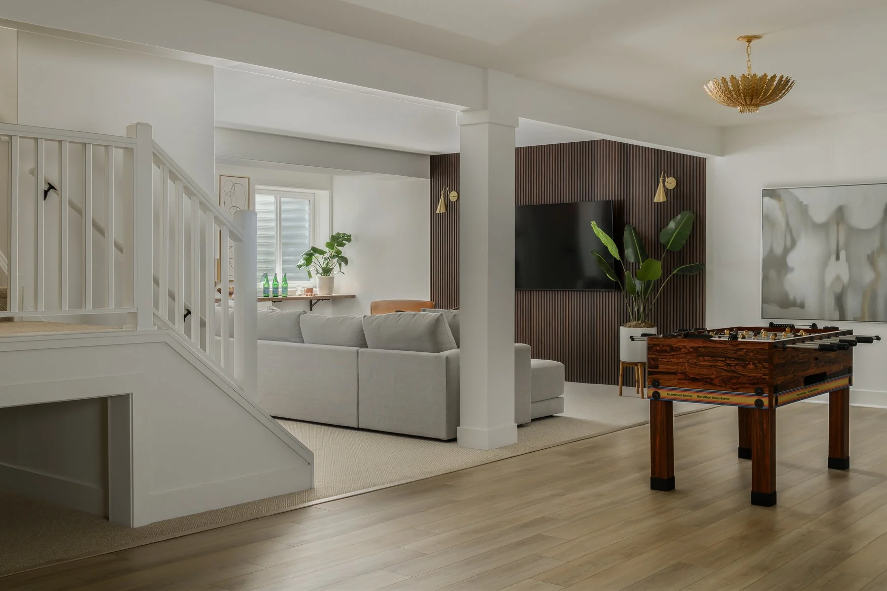 Living room with white walls, a white staircase, a beige sofa, a TV mounted on a wooden-paneled wall, a large abstract painting, a potted plant, a foosball table, and natural light coming through a window.