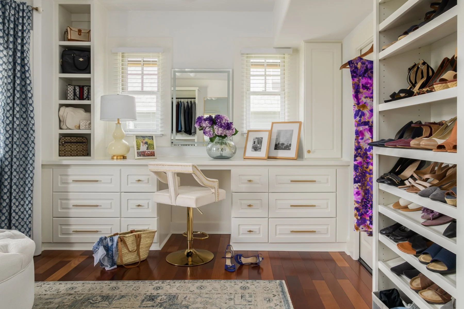 A walk-in closet with white built-in drawers and shelves, a large mirror, a white swivel chair, a table with a flower vase and picture frames, a purple floral curtain, and a shoe rack filled with various shoes.