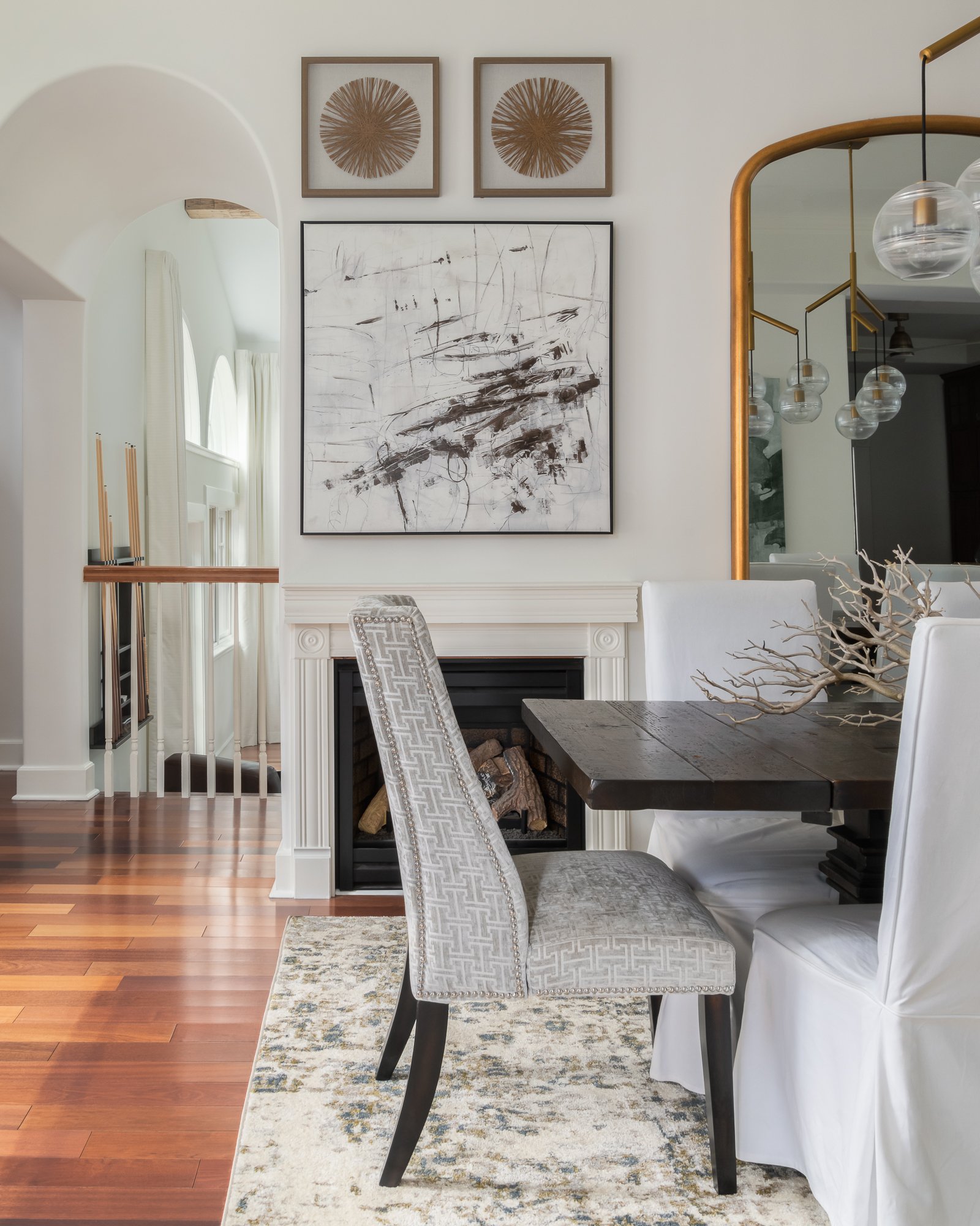 Dining area with black wood table, white and upholstered chairs, and a fireplace with artwork above it. Decorative items include a branch centerpiece and framed wall art.