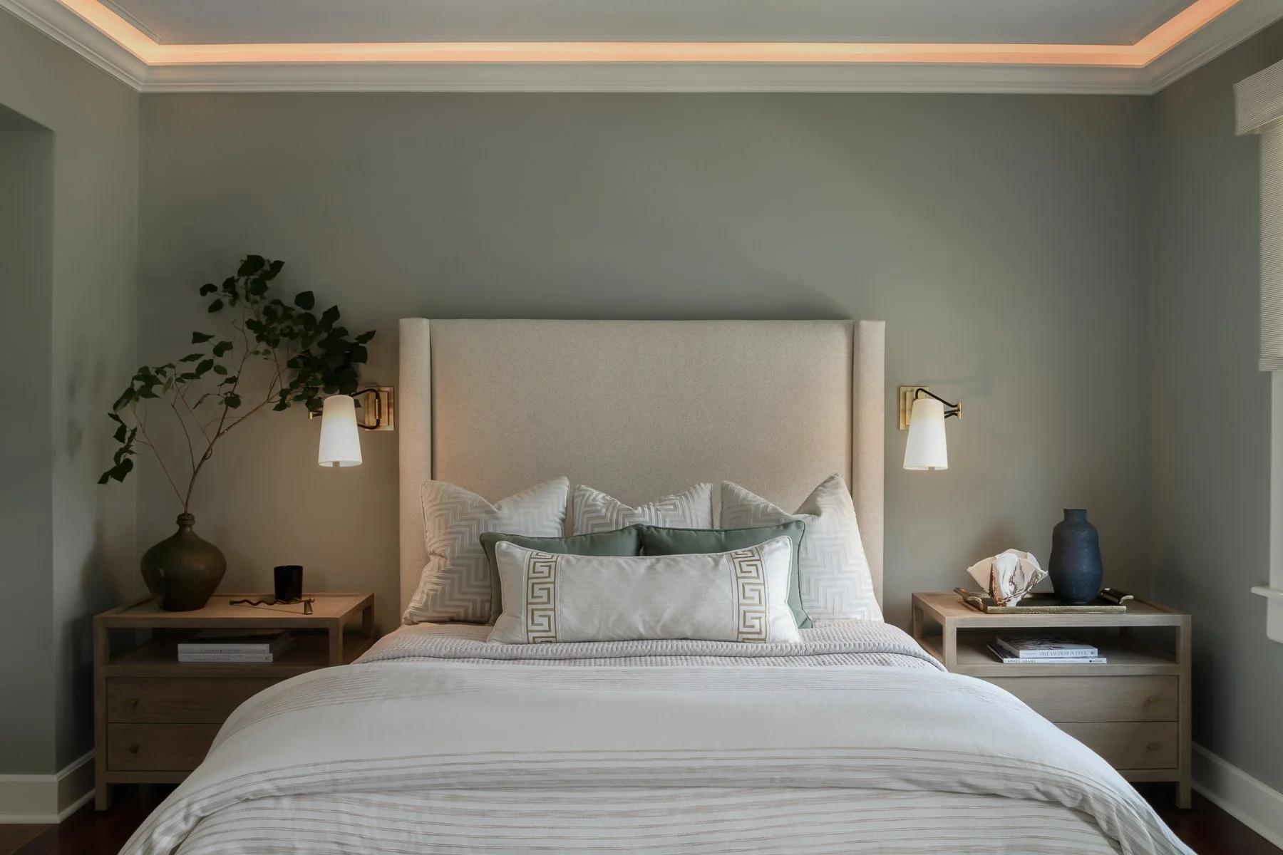 A neatly made bed with white and green pillows, beige headboard, green walls, and two wooden nightstands with decorative items and lamps, in a modern bedroom.
