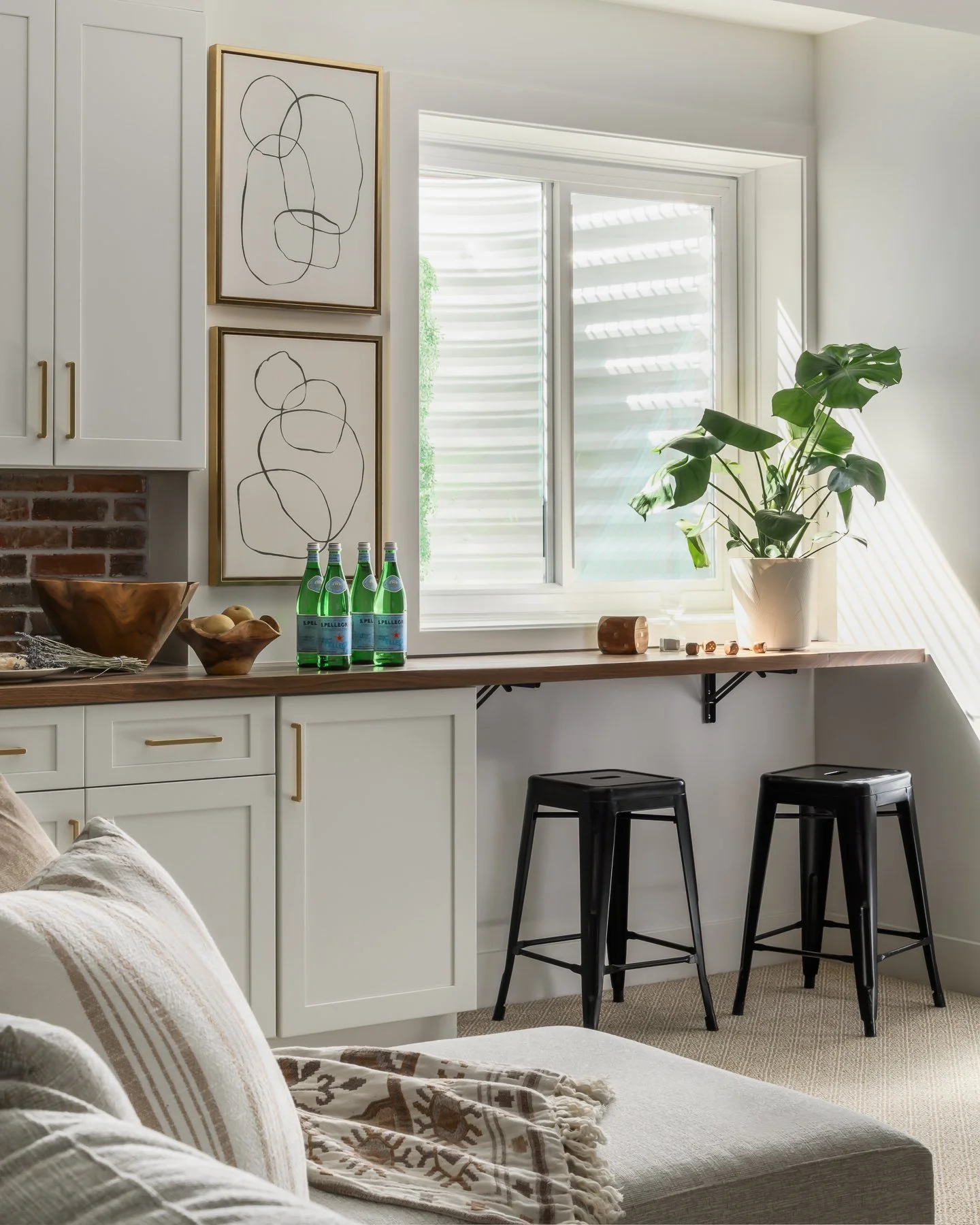 Close-up view of a living room with a beige sofa, a throw pillow, and a blanket. In the background, there's a kitchen counter with decorative wooden bowls, four glass bottles of sparkling water, and two black bar stools. The wall has white cabinetry,
