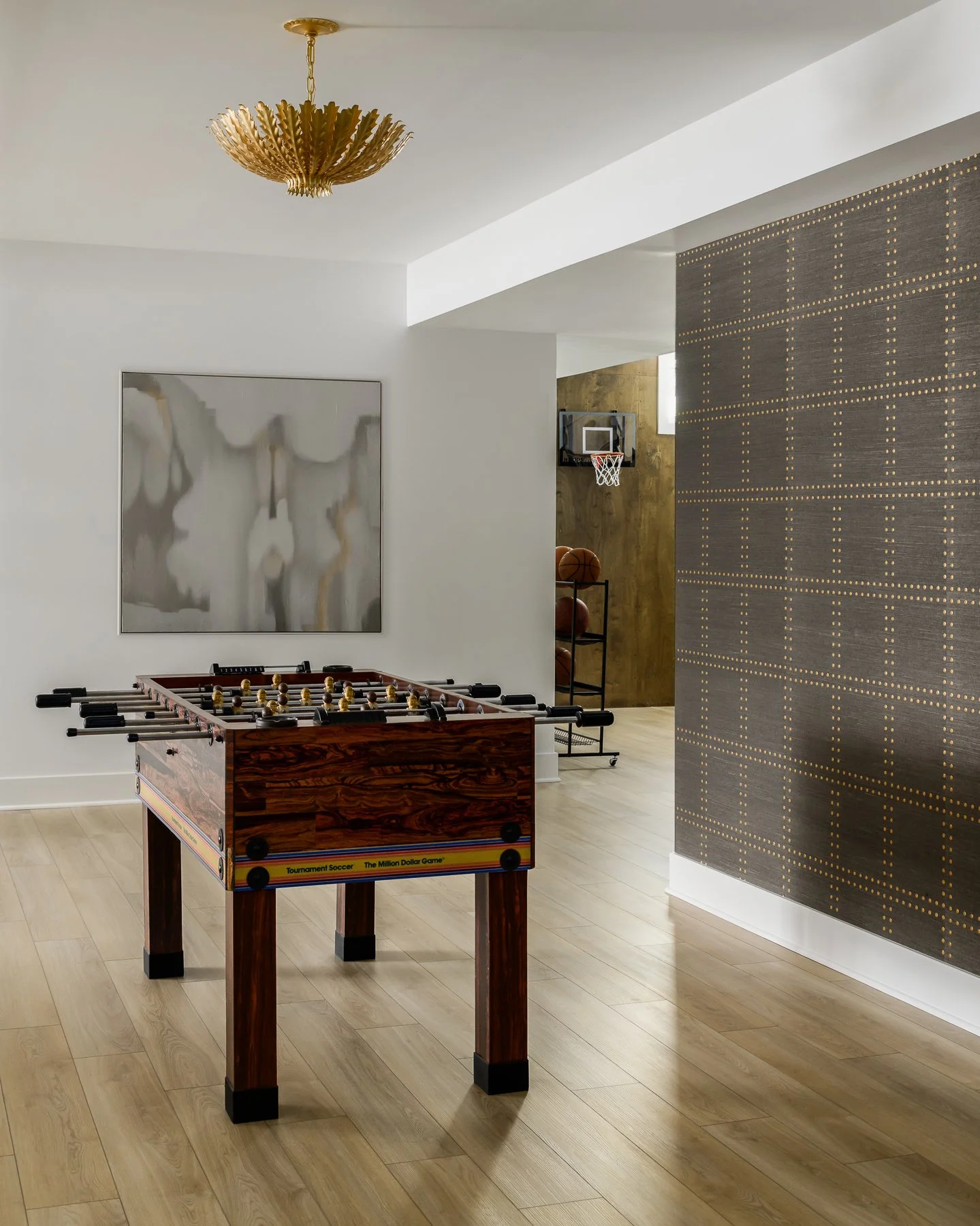 A room with wooden flooring, featuring a foosball table in the foreground, a large abstract painting on the white wall, a wall-mounted basketball hoop in the background, and a decorative wall with a grid of small lights.