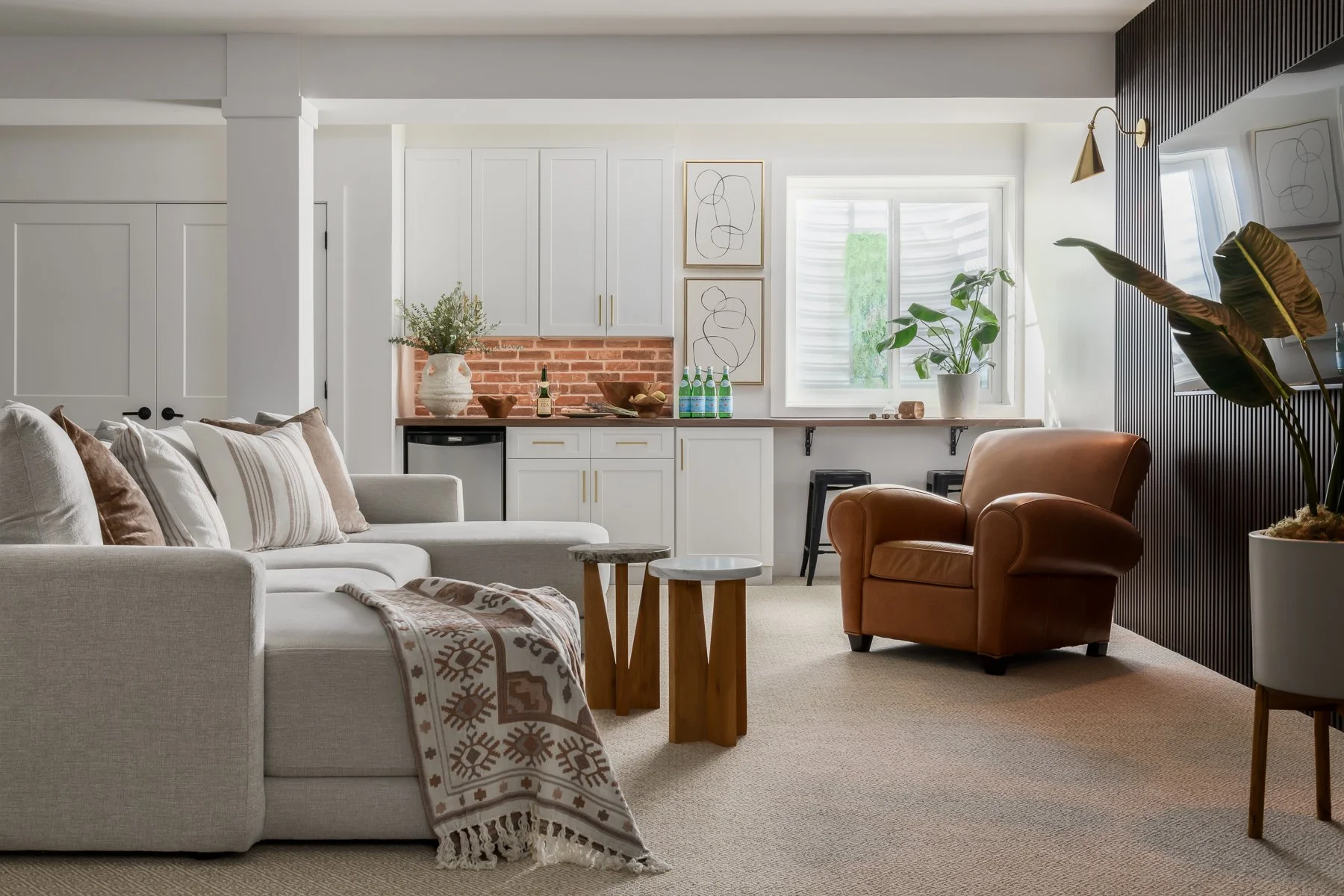 Living room with white sofa, patterned throw blanket, small tables, brown leather armchair, wall art, large window with blinds, potted plants, and a kitchen in the background with white cabinets, brick backsplash, and bar stools.
