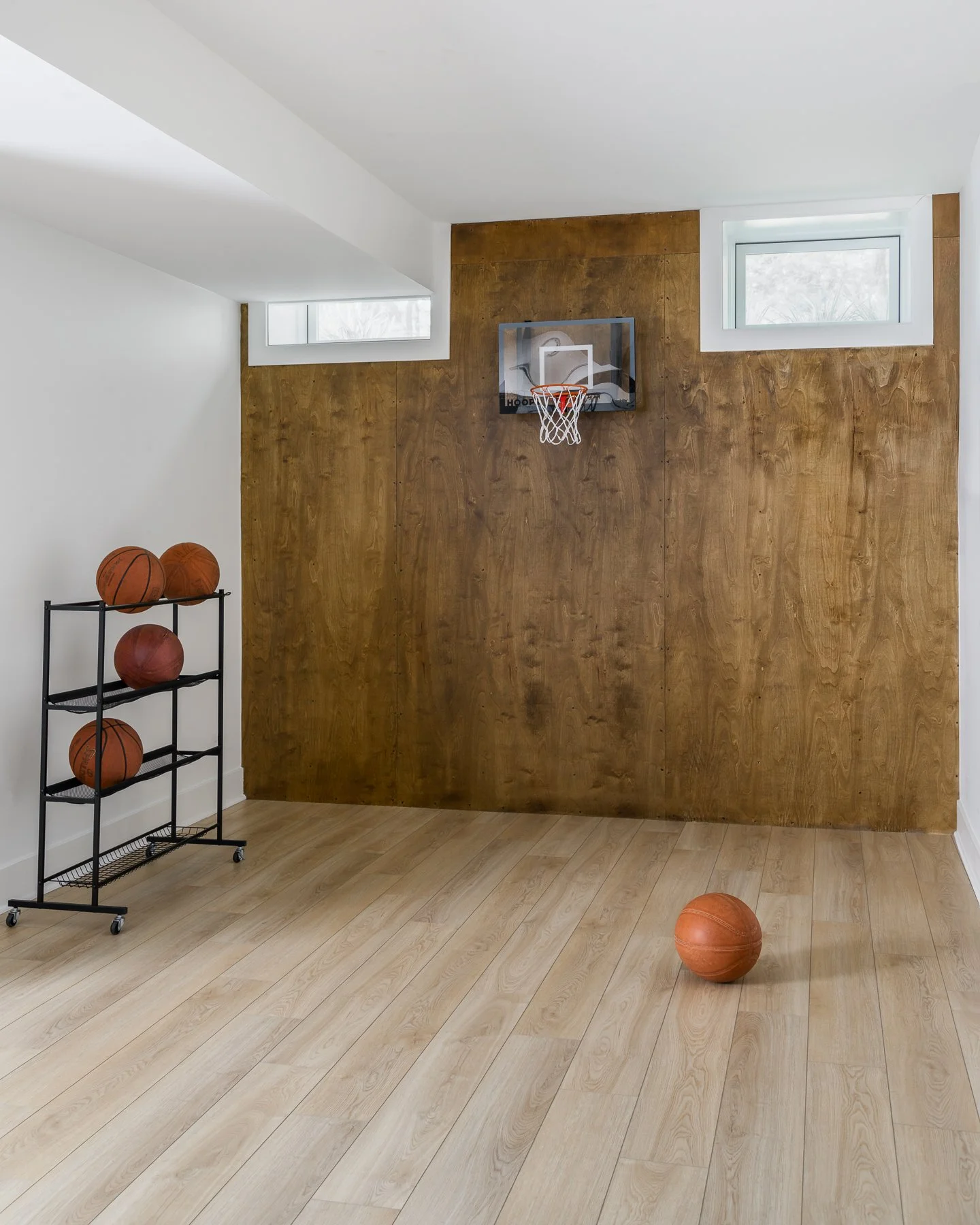 Indoor basketball practice area with a wall-mounted hoop, a small wheeled rack holding basketballs, and a basketball on the wooden floor.