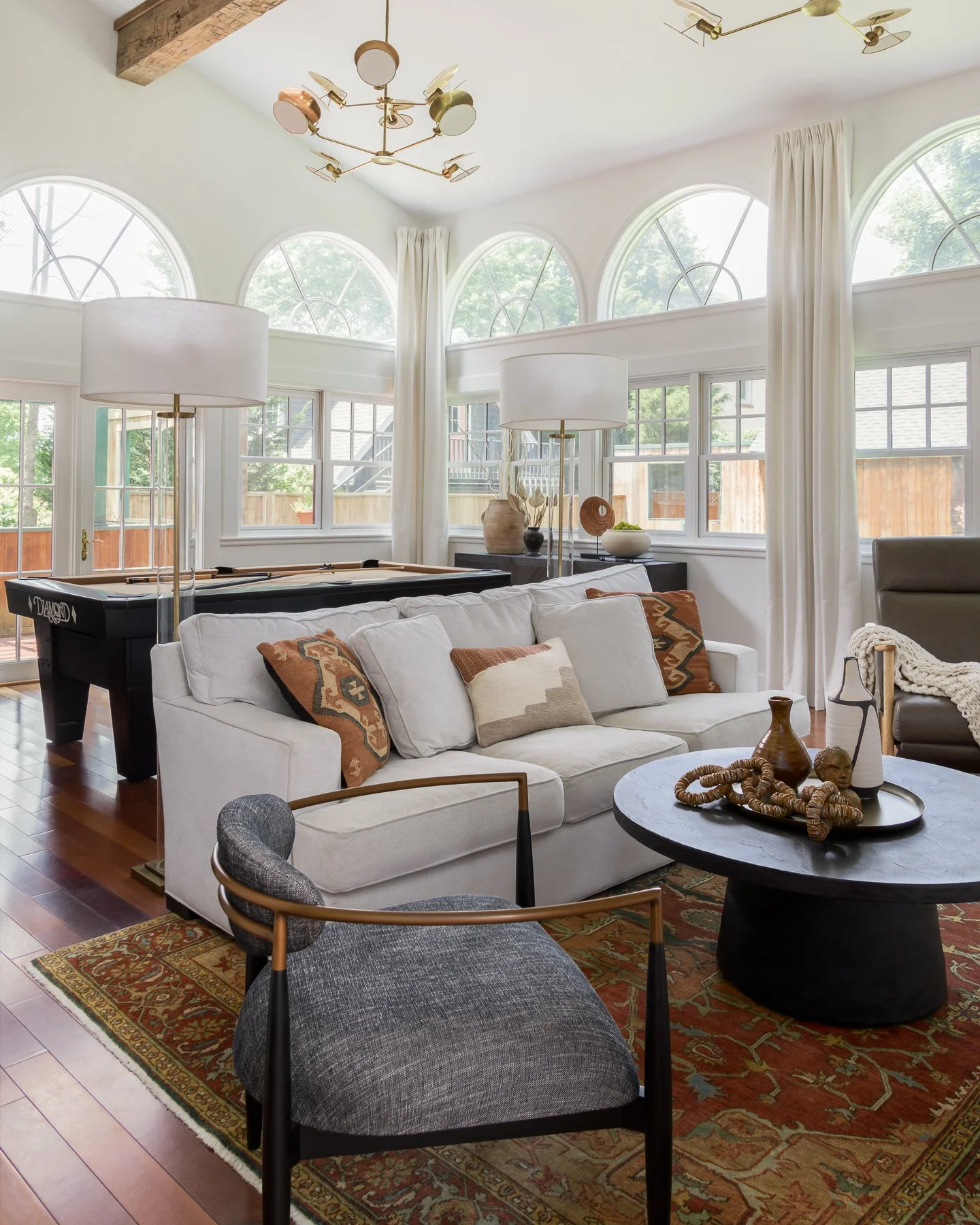 A living room with white walls and large arched windows letting in natural light. It has a gray armchair, a white sofa with patterned throw pillows, a round black coffee table with decorative items, two tall white lamps, a vintage rug, and a pool tab
