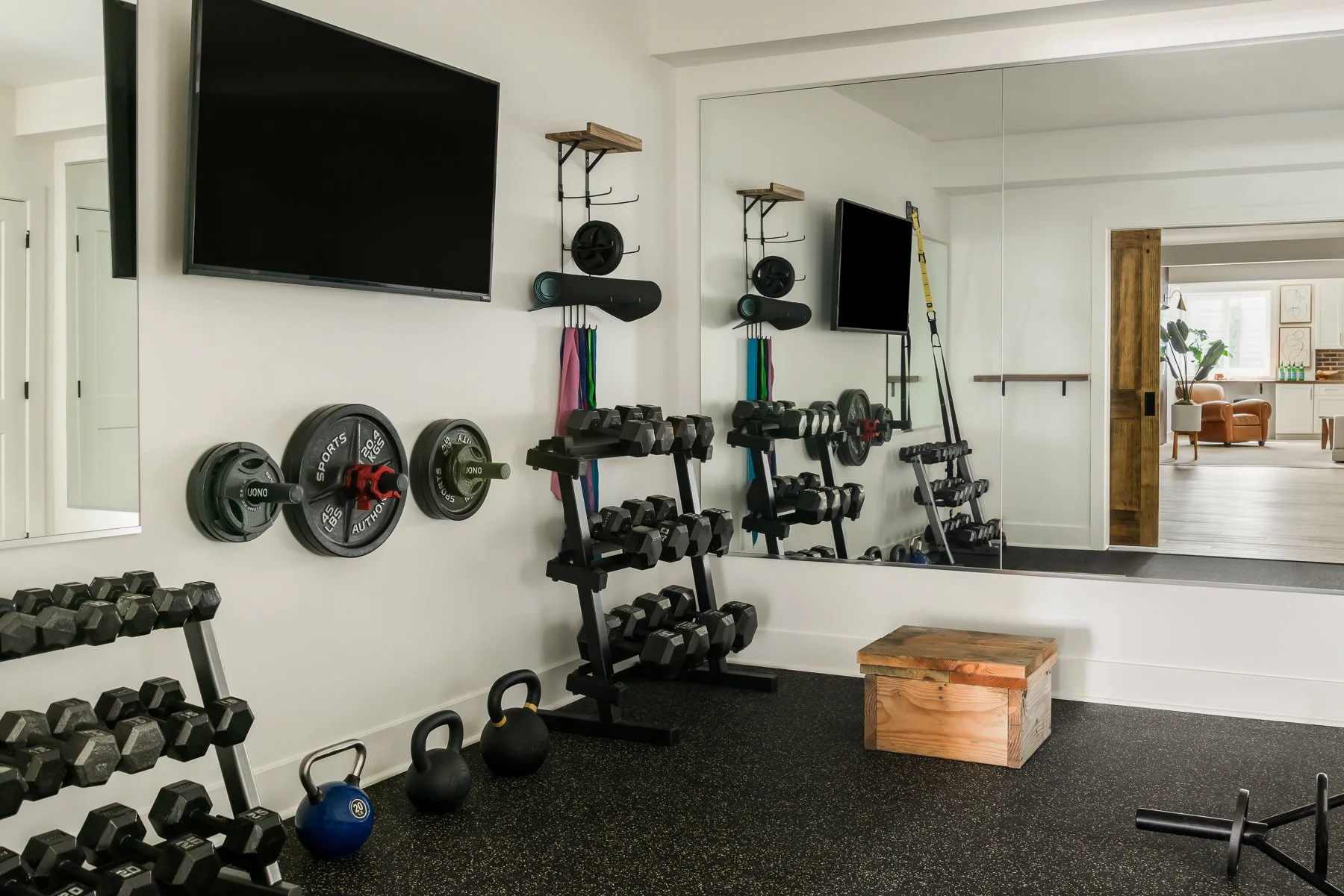 A home gym with wall-mounted TV, dumbbell racks, kettlebells, resistance bands, and a large mirror. There is a wooden block and a wall with hooks for workout equipment. The room opens into a living space with a chair, plant, and framed artwork.