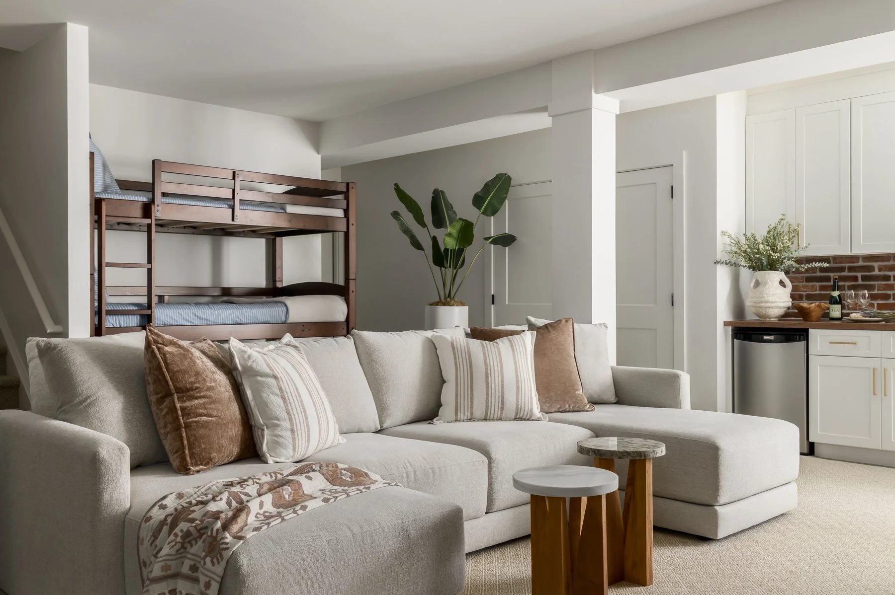 A cozy living room with a beige sectional sofa adorned with brown and striped pillows. Behind the sofa, there's a large potted plant with broad green leaves. In the background, white cabinets and a small mini-fridge are visible, along with a brick ba