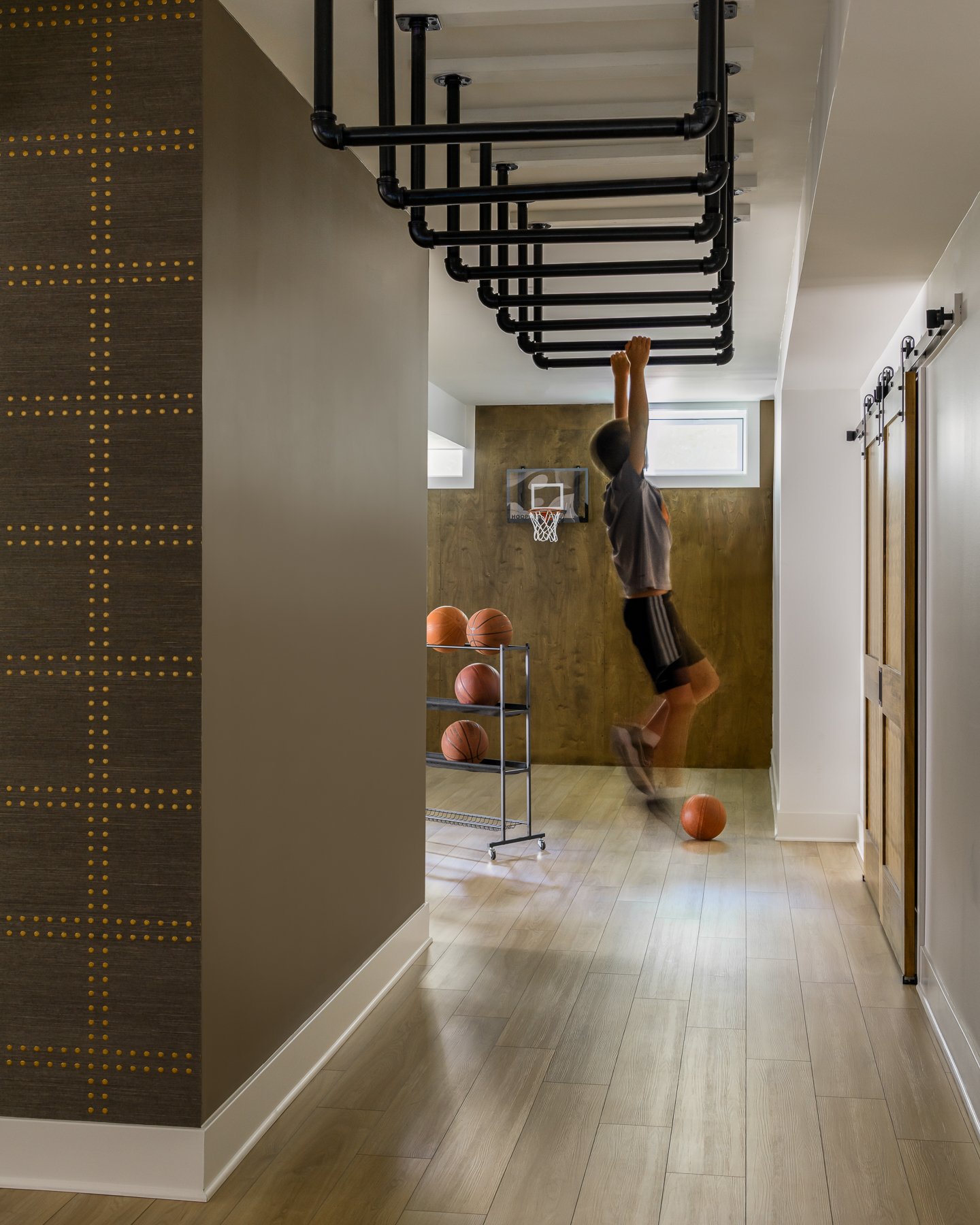A young boy playing basketball indoors in a room with wooden flooring and a small basketball hoop. He's jumping to make a shot, with a basketball on the floor near him and a rolling cart of basketballs nearby. The room has a window and a decorative w