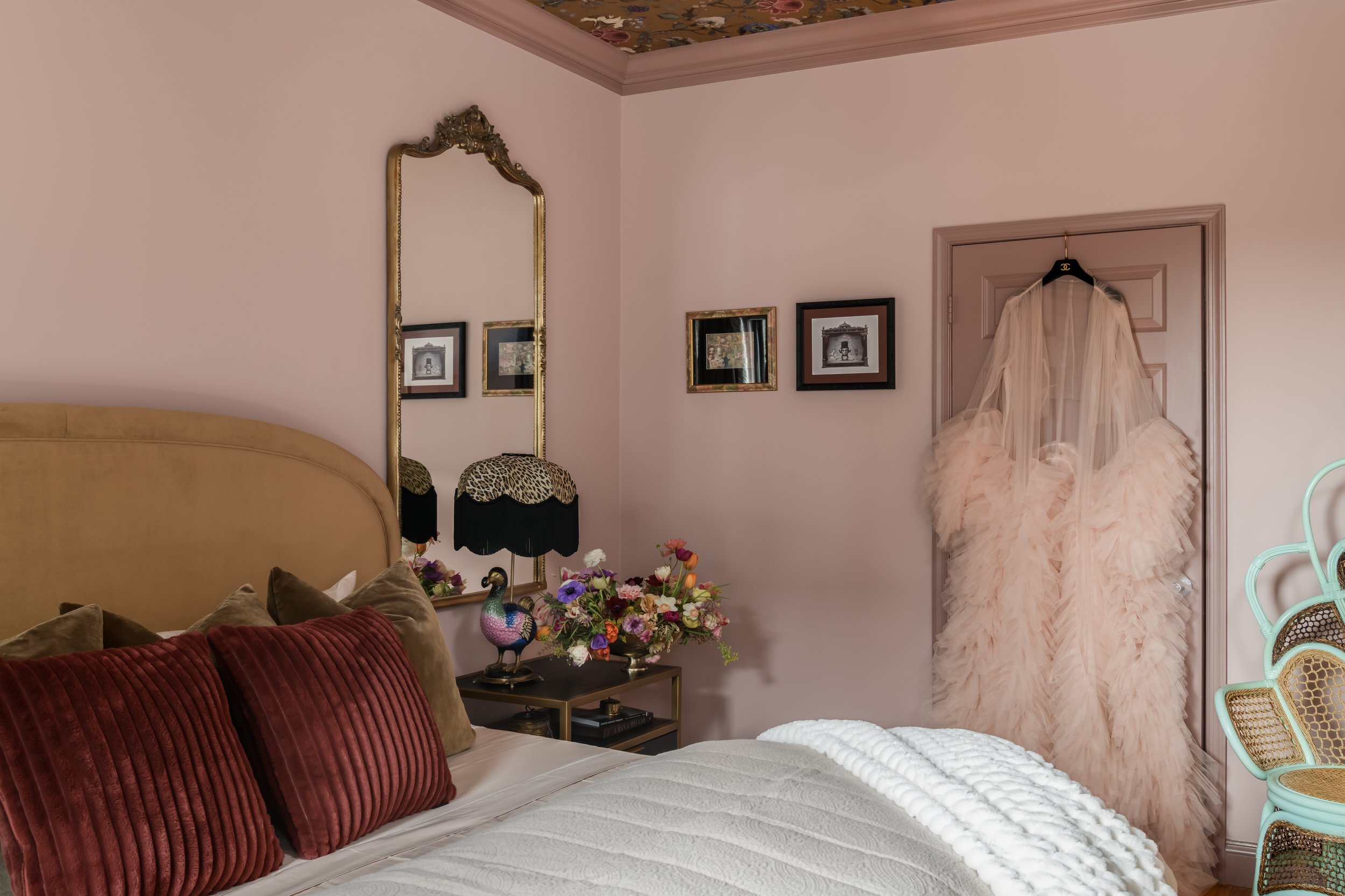 A cozy bedroom with a bed, adorned with velvet pillows and a textured white bedspread. Beside the bed, there's a side table with a colorful peacock lamp and a floral arrangement. The wall features framed artworks, a tall mirror with an ornate gold fr